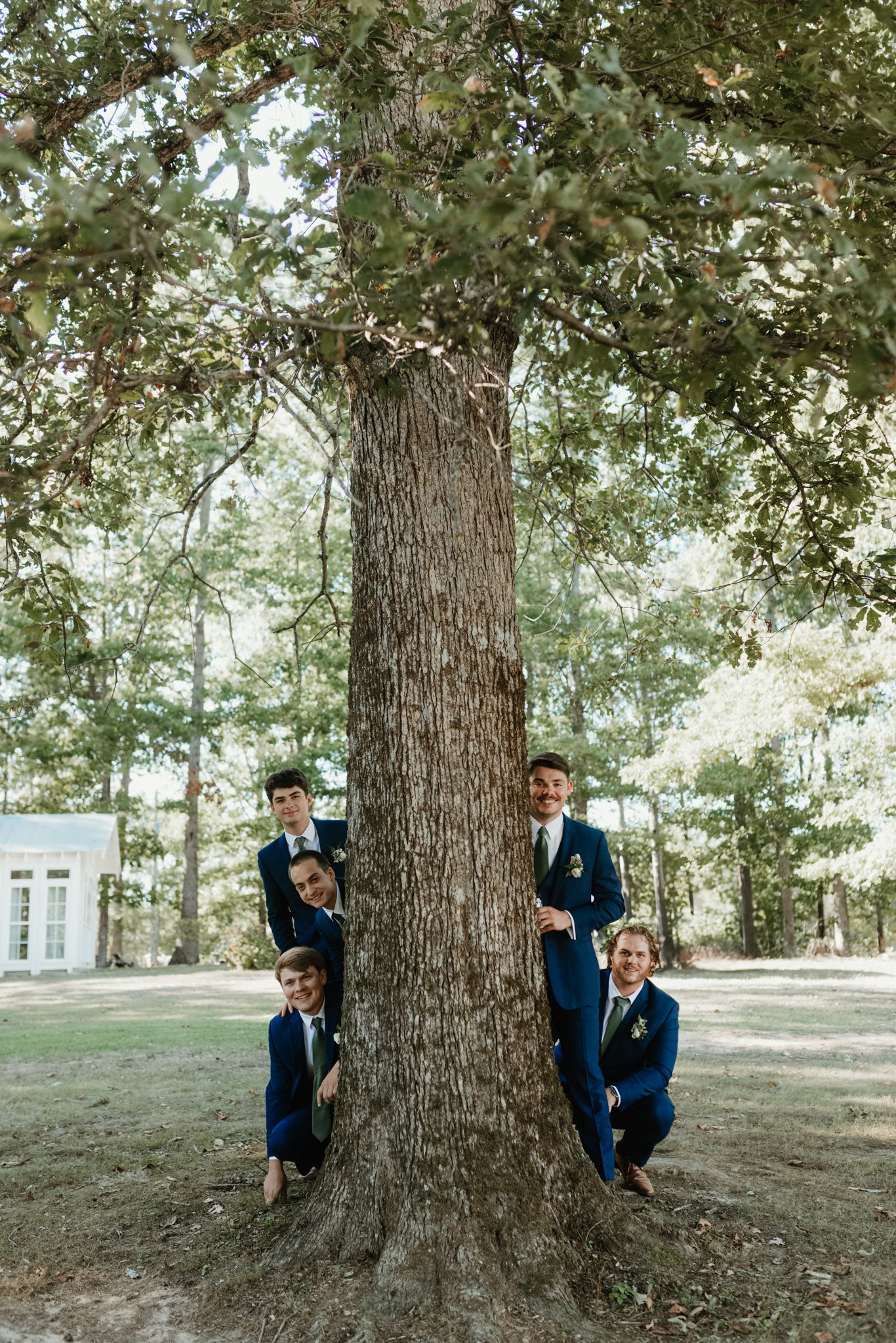 Four figures in matching navy suits pose playfully around a large tree trunk in an outdoor woodland setting.