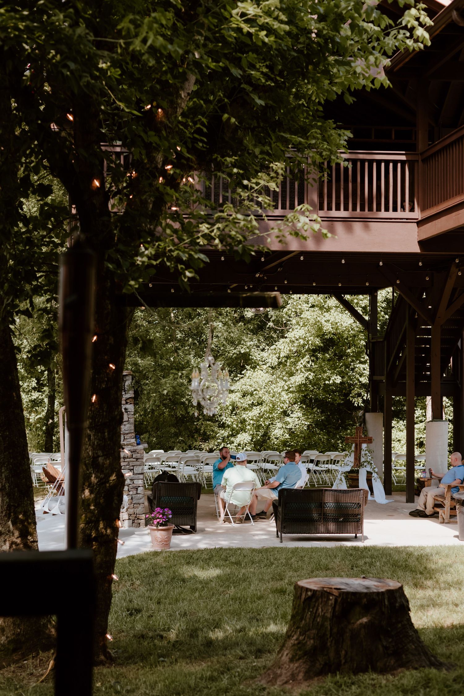 Outdoor wedding reception setup in a rustic wooded area with string lights, wooden deck above, and tables for guests.