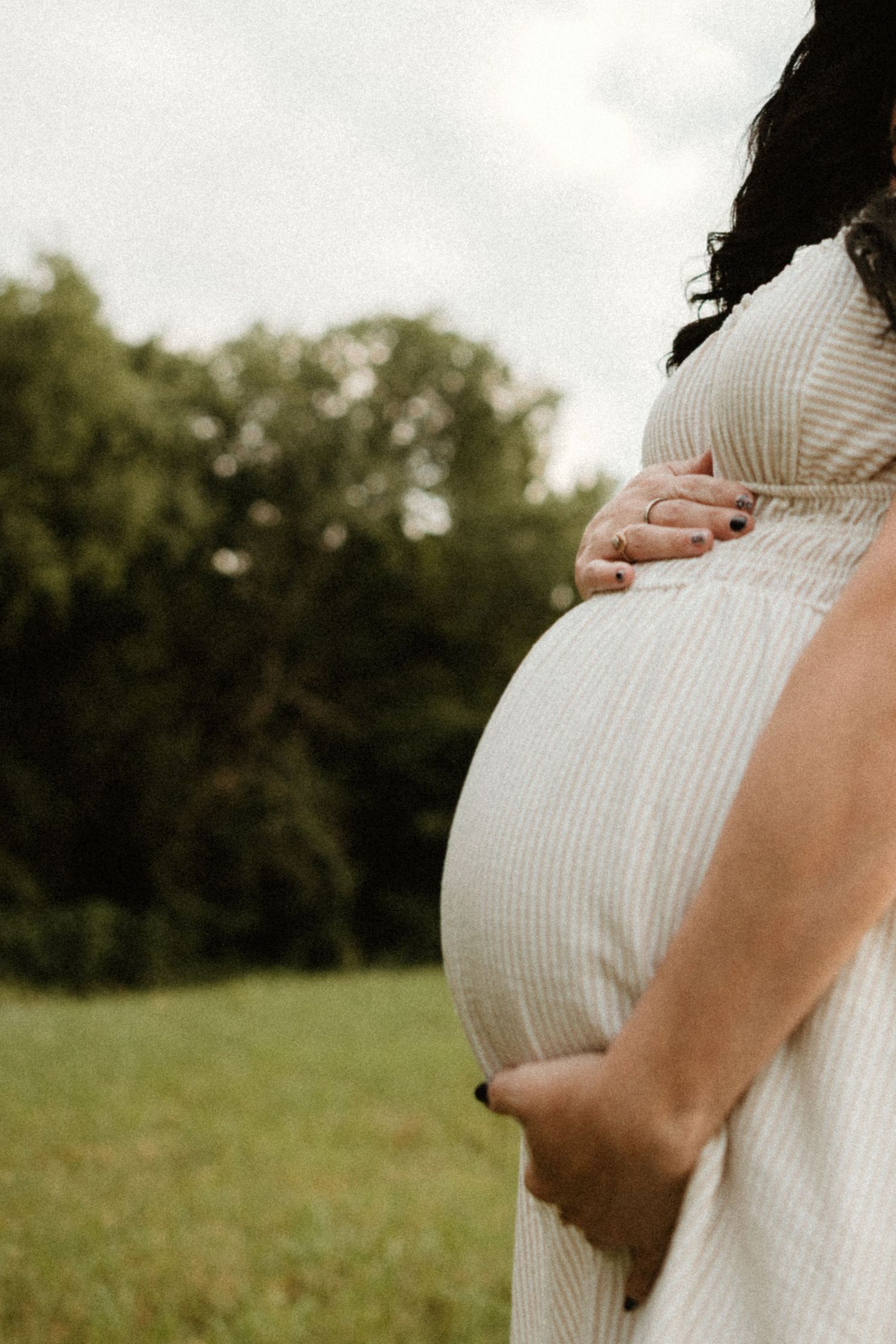 Person wearing a white dress cradles their pregnant belly while standing in a grassy field on an overcast day.