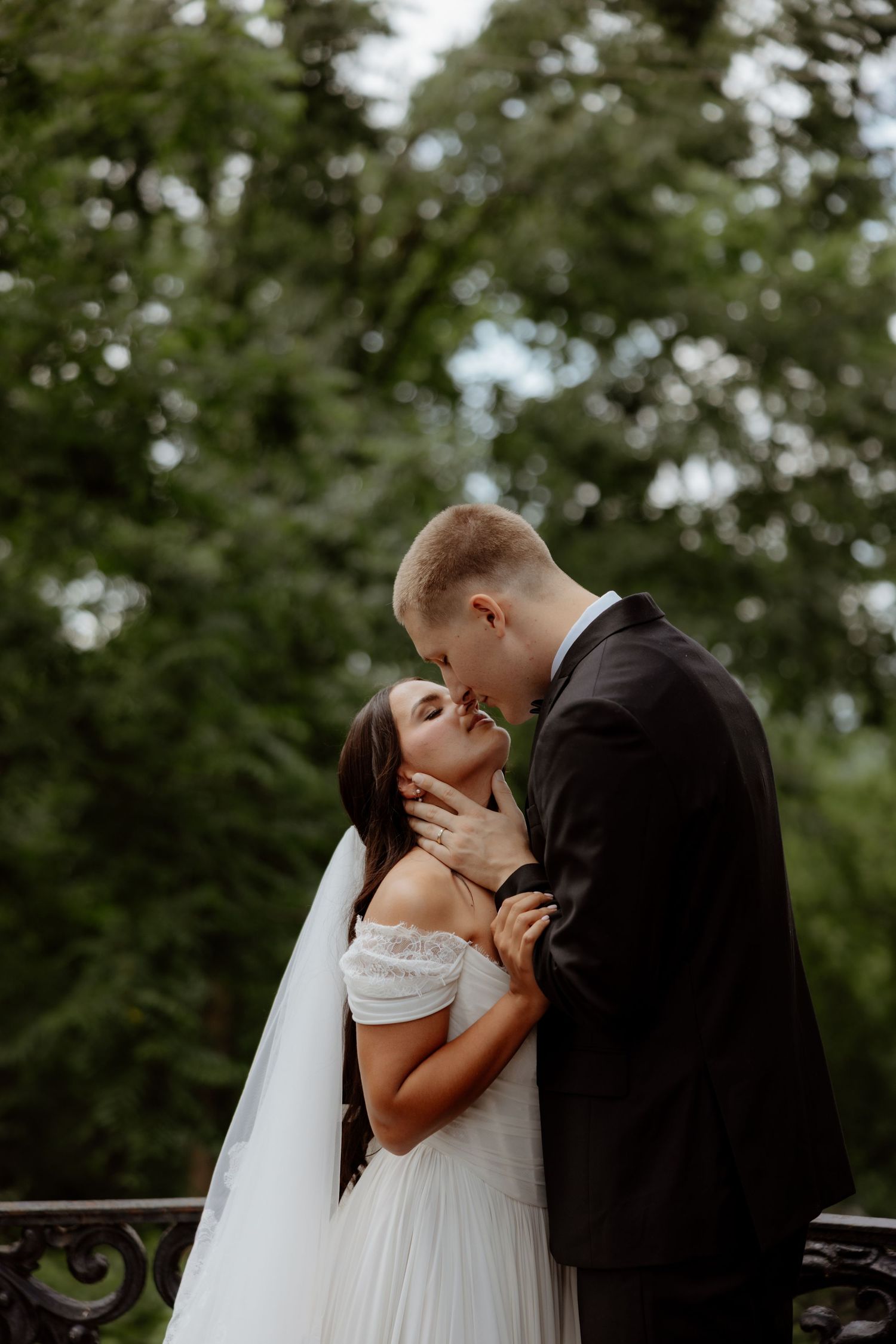 Bride and groom share a romantic kiss outdoors, surrounded by lush green trees, bride wearing off-shoulder gown and veil.