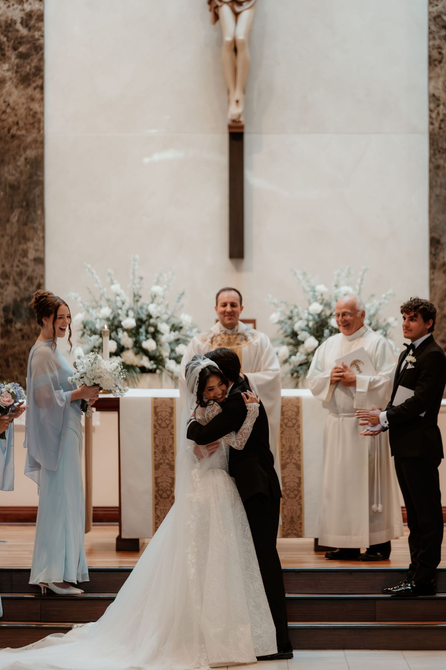 Bride and groom share first kiss at church altar with smiling officiant and wedding party nearby.