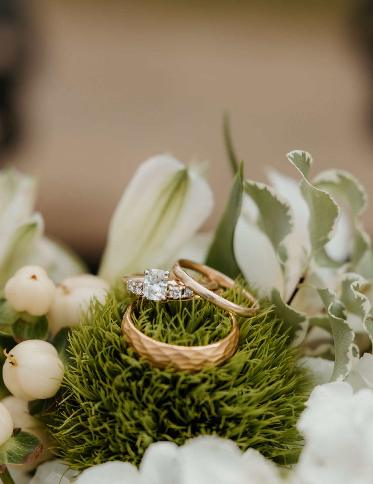 Gold wedding bands and diamond engagement ring nestled in green moss surrounded by white flowers and berries.