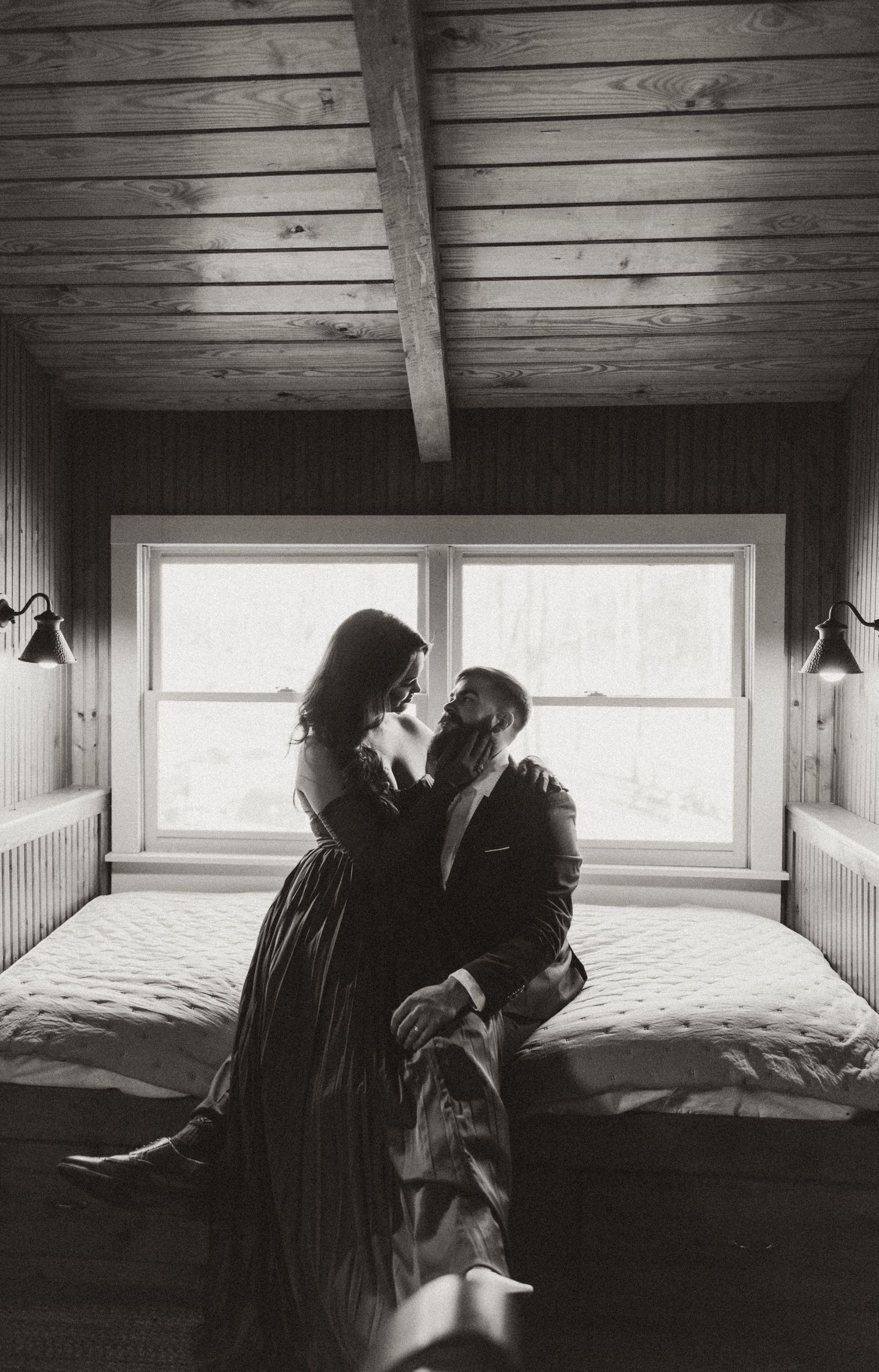Black and white photo of a couple sharing a romantic moment on a bed in a rustic wood-paneled cabin room.