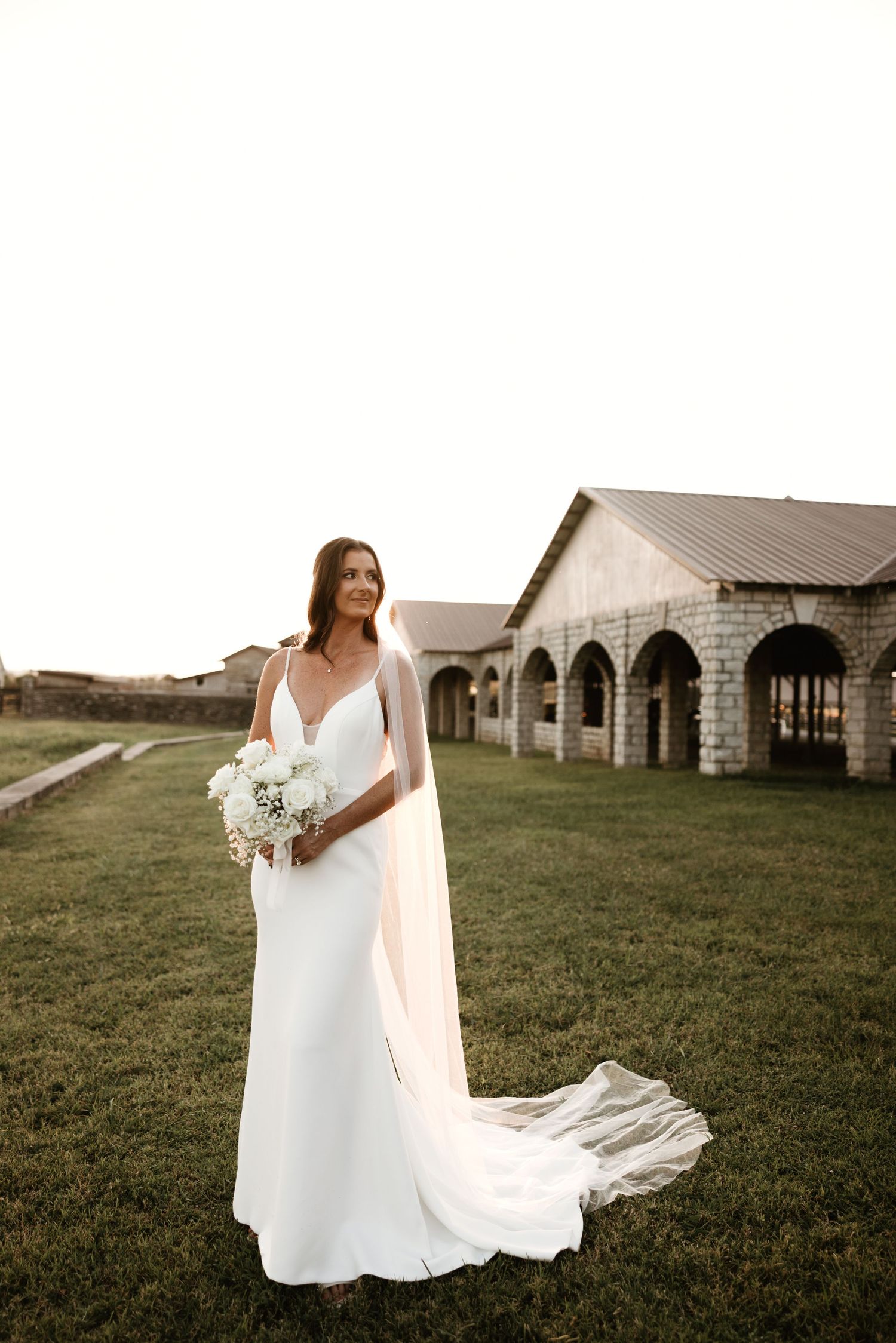Bride in flowing white dress poses elegantly near rustic stone buildings in a countryside setting.