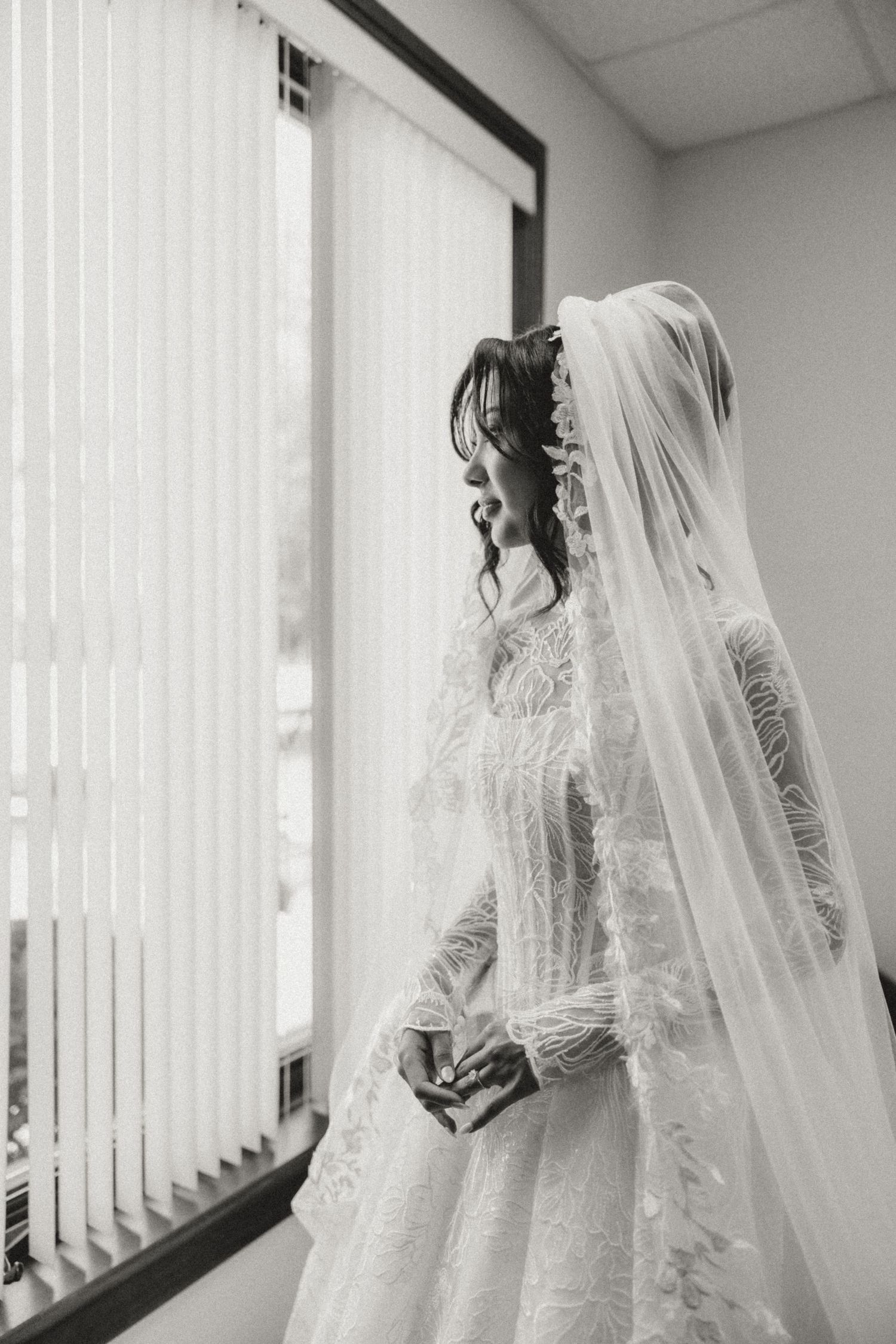 Elegant bride in lace long-sleeve gown and cathedral veil stands by window in black and white photo.