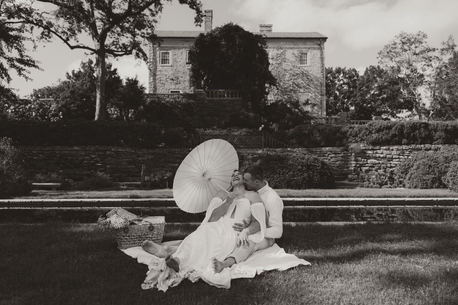 Black and white photo of a bride and groom sitting on grass with a parasol in front of an elegant stone mansion.