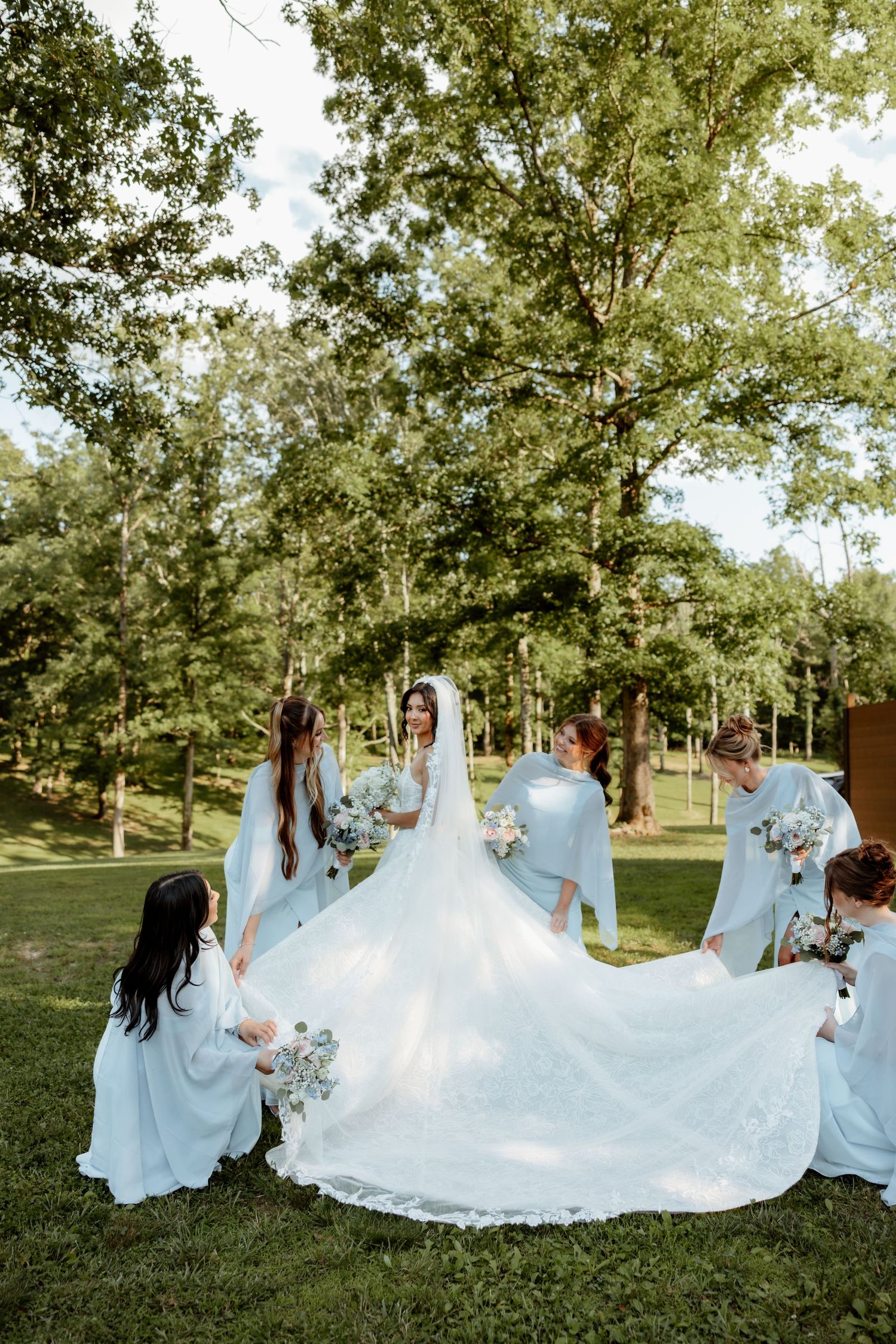 Bridesmaids in light blue dresses arrange bride's white cathedral train on green grass outdoors.