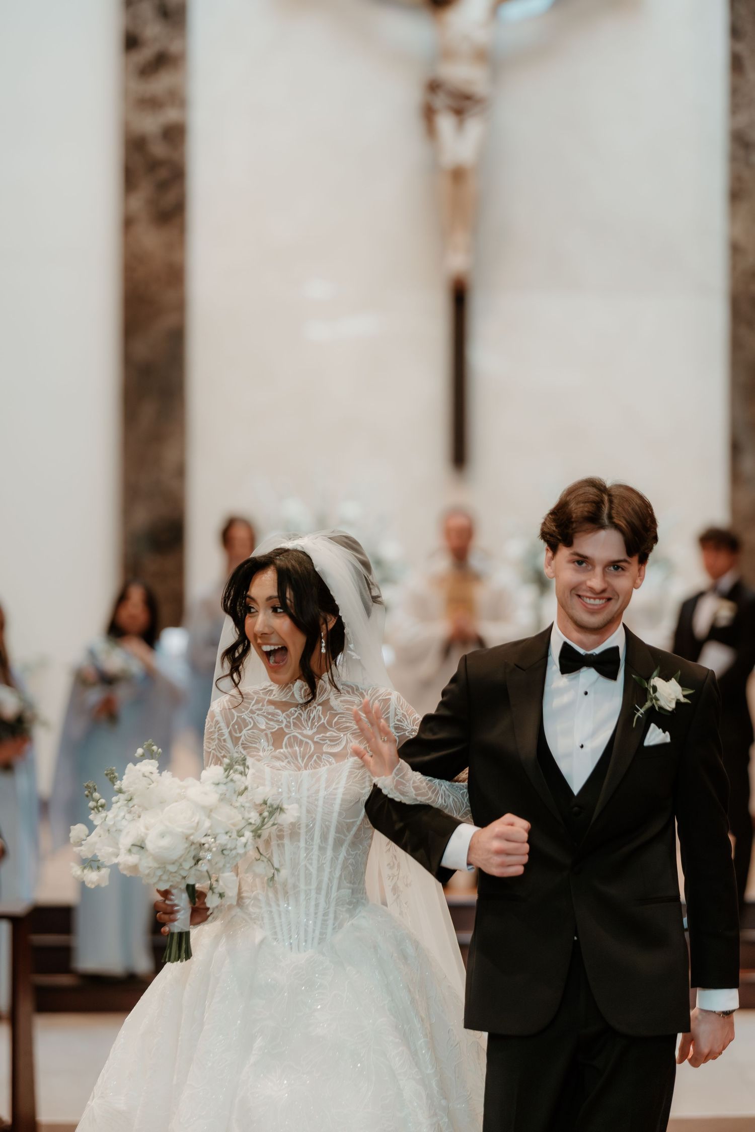Joyful bride in lace gown and groom in black tuxedo walking down church aisle after wedding ceremony.