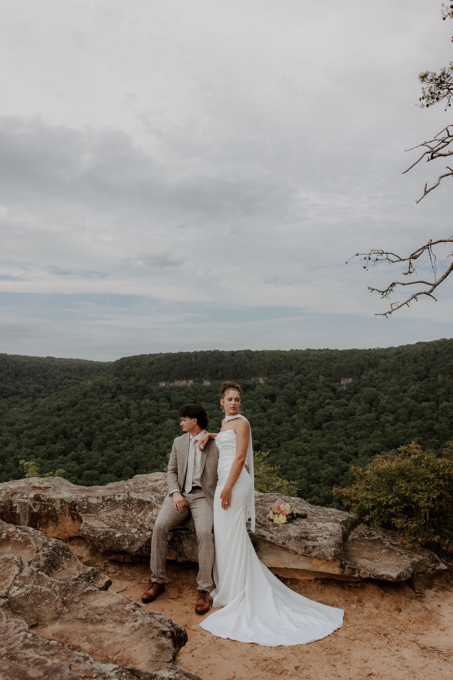 Bride and groom pose on rocky cliff overlook with lush green forest valley backdrop on their wedding day.