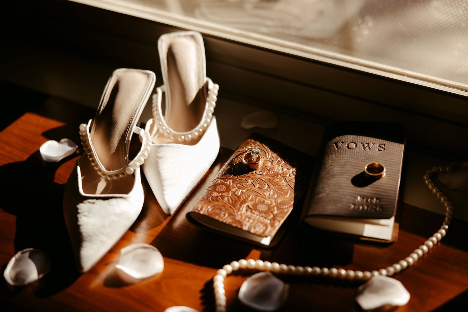 White pearl bridal heels, gold clutch, wedding vows book, and pearl necklace arranged on a wooden windowsill.