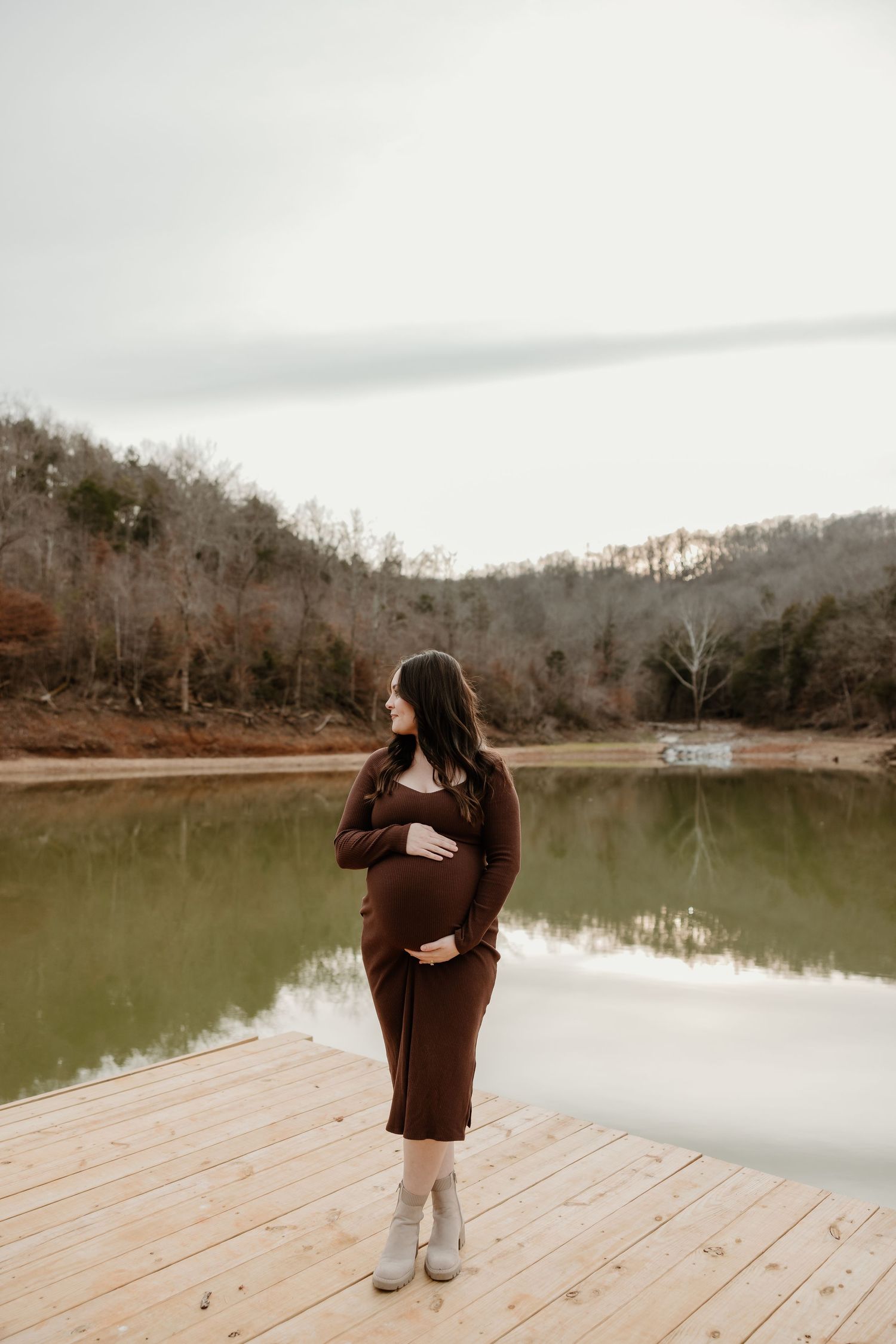 Pregnant woman in brown dress on wooden dock by reflective lake with autumn hills backdrop.
