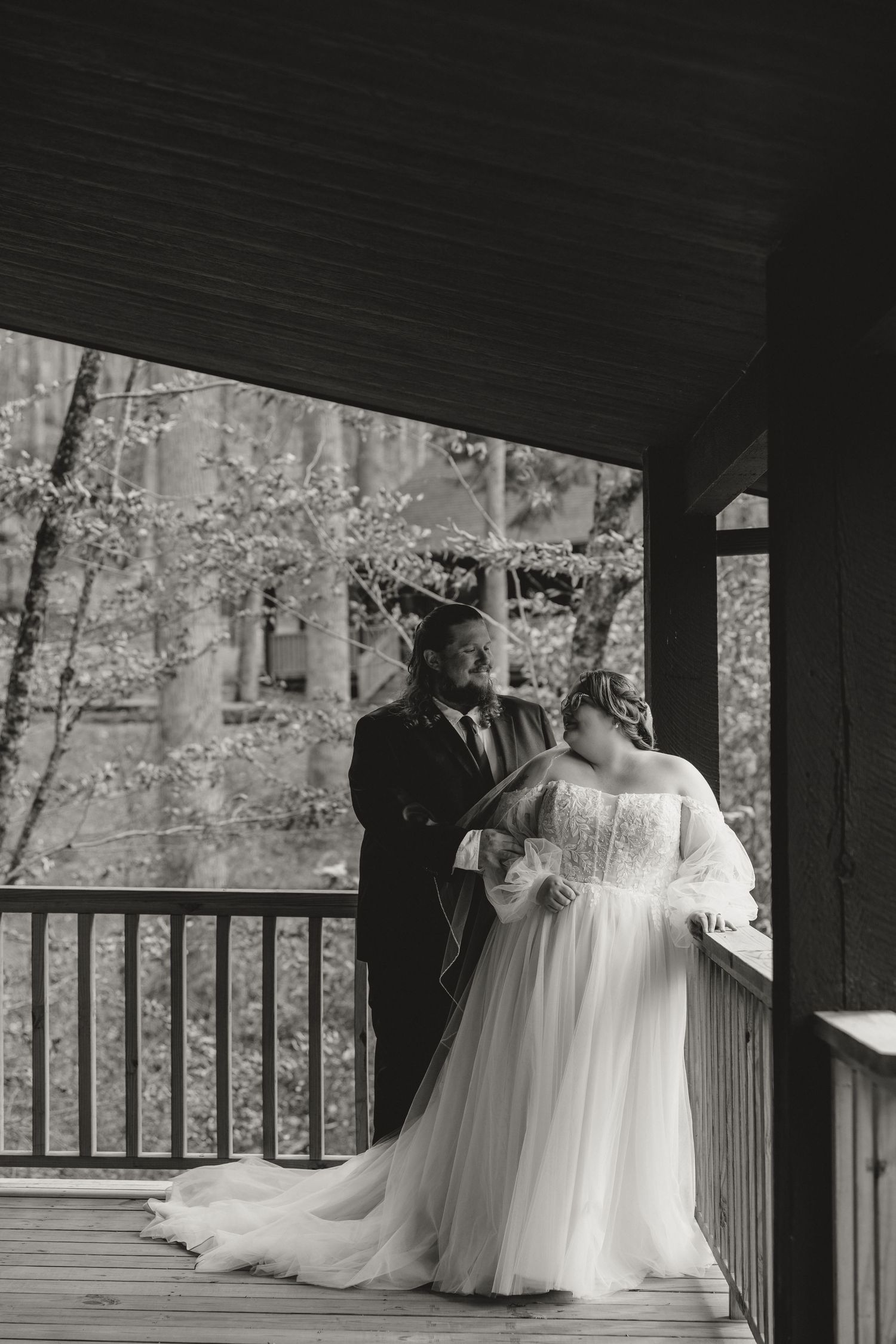 Black and white photo of bride and groom sharing a tender moment on a wooden porch surrounded by blooming trees.