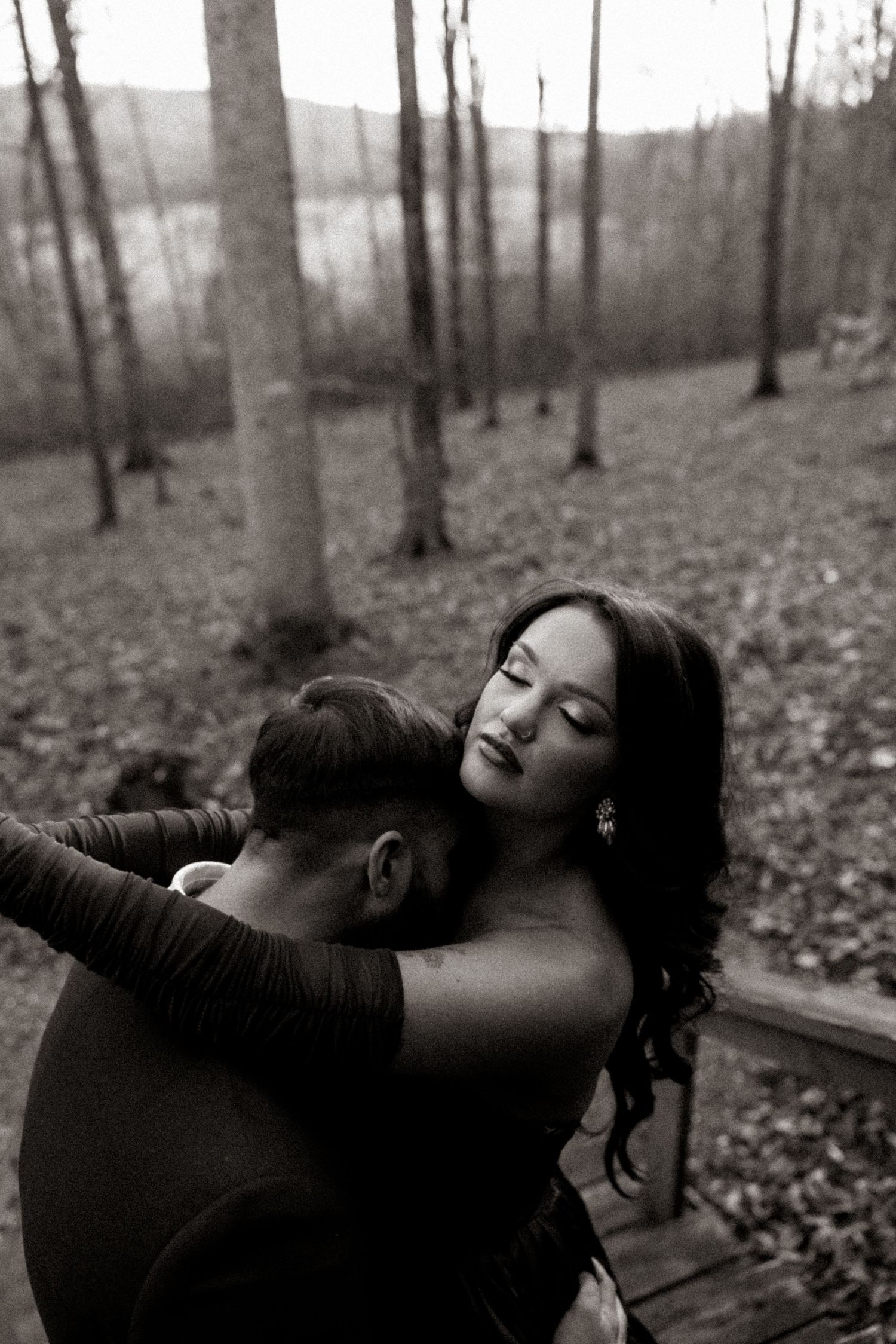 Black and white photo of a couple embracing in a forest, woman with eyes closed leaning on mans shoulder.