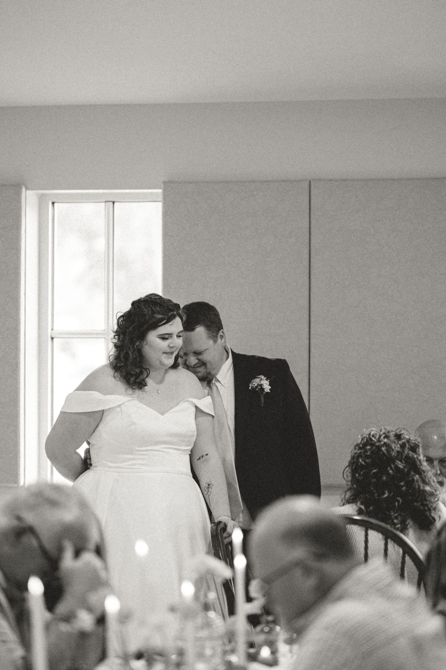 A couple shares an intimate moment during their wedding reception as guests look on in black and white photography.