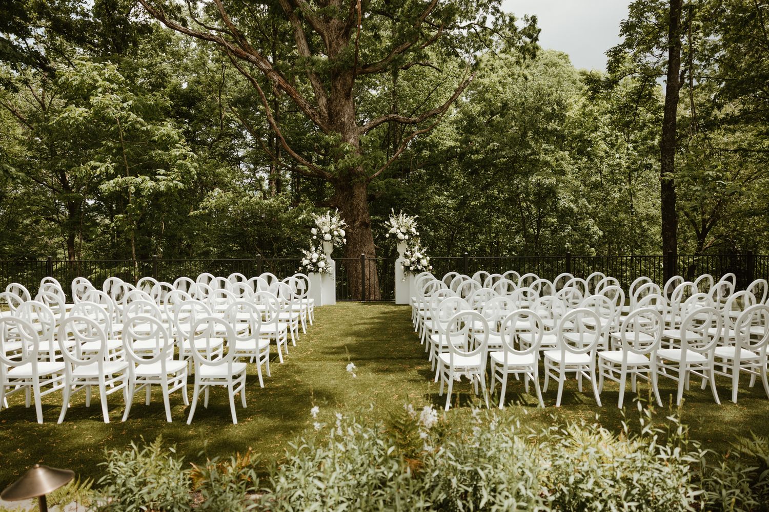White chairs arranged in rows for an outdoor ceremony surrounded by trees and greenery.