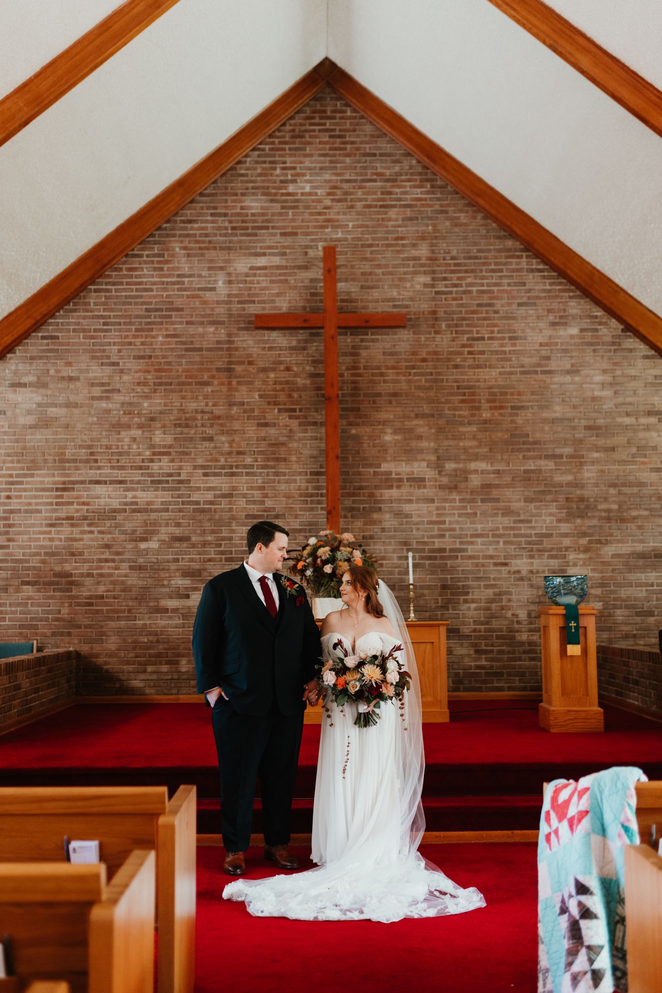 A traditional church wedding ceremony with a wooden cross on a brick wall and red carpet in the aisle.