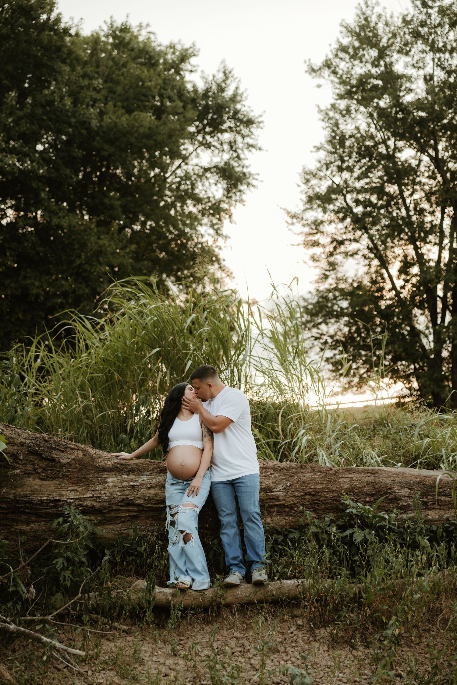 A serene natural outdoor photo session features two people in light colored clothing standing by fallen tree branches at sunset.