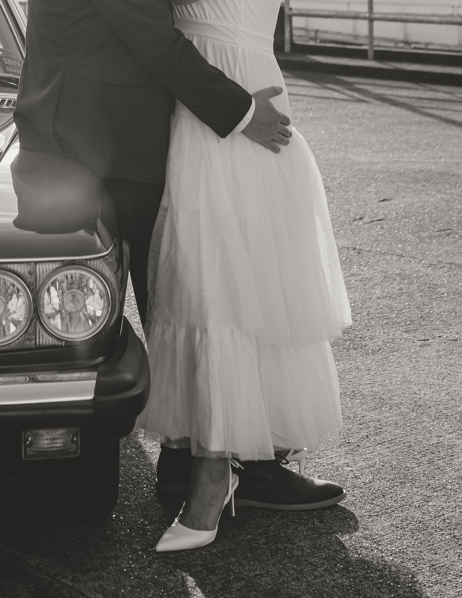 Black and white photo showing dress shoes and flowy white gown next to a classic car's front headlight.
