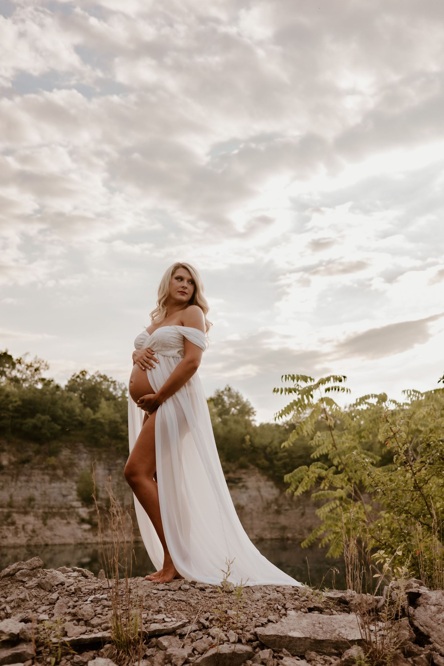 A maternity photo shoot features an elegant white off-shoulder dress against a dramatic cloudy sky and rocky terrain.