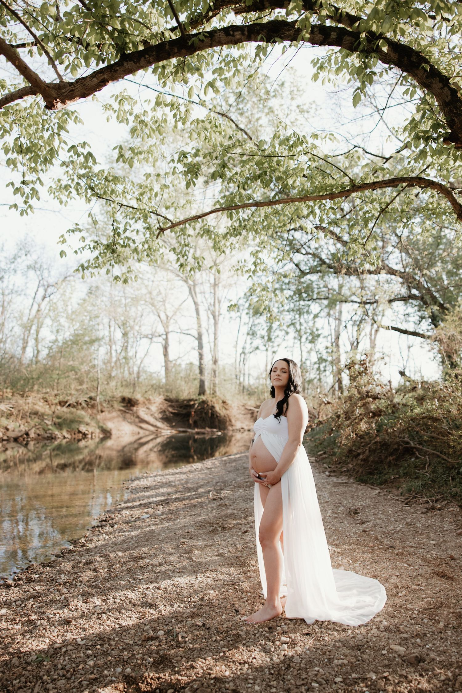 A dreamy maternity photo by a creek with flowing white dress and natural sunlight.
