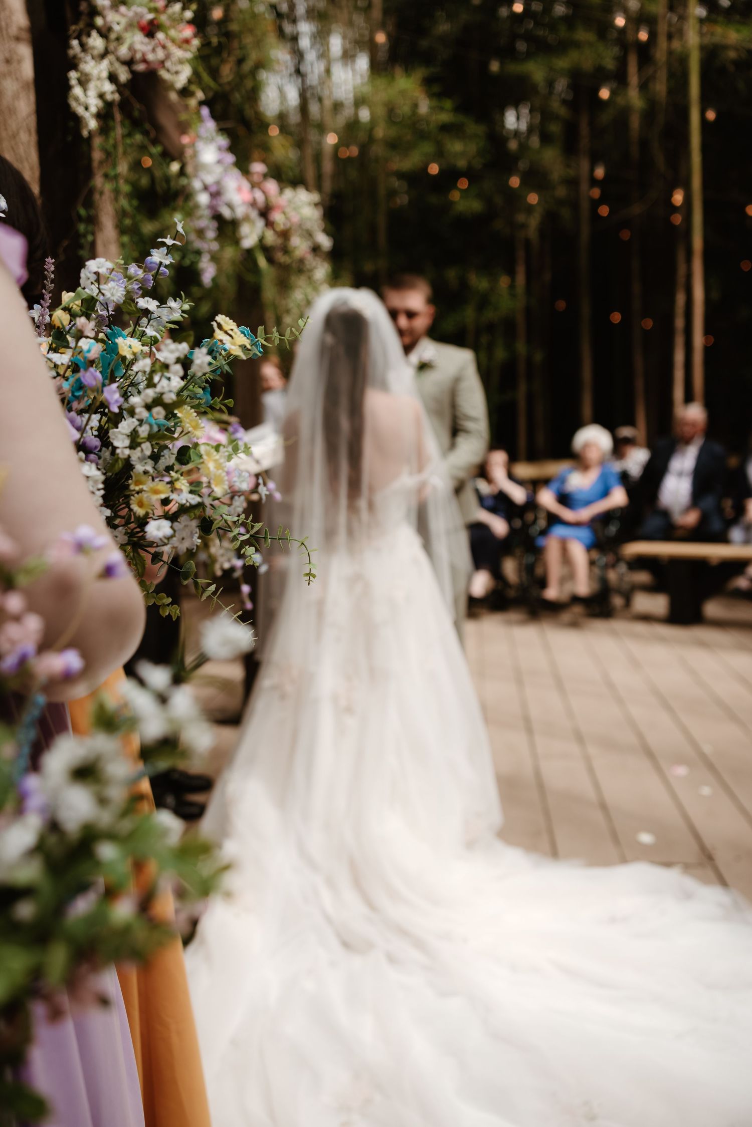 A scenic outdoor wedding ceremony with floral decorations and flowing white dress train in focus.