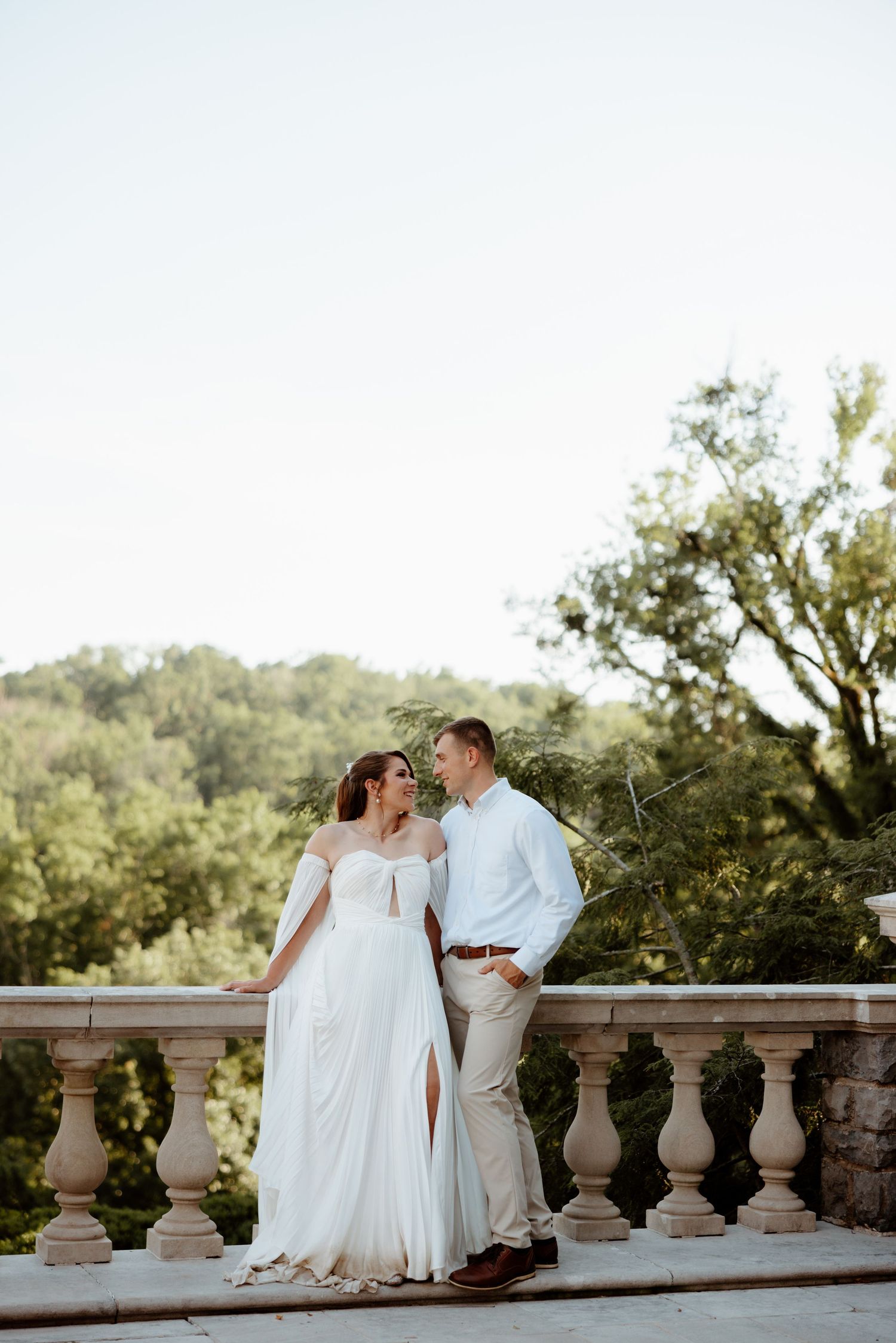 Bride and groom share a romantic moment on a stone balcony overlooking lush green trees on their wedding day.