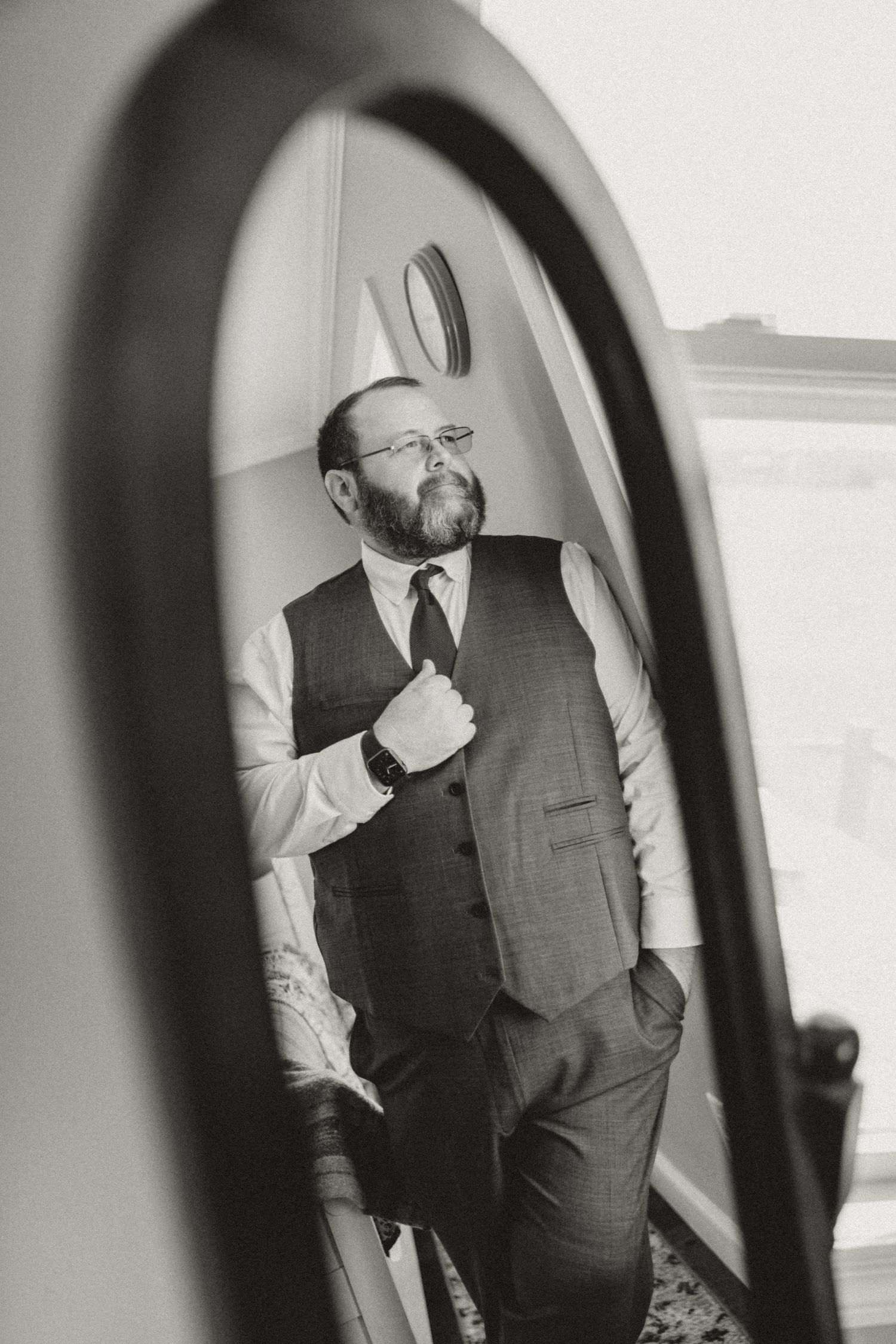 Black and white mirror reflection of a bearded groom in vest and tie adjusting his outfit before wedding ceremony.