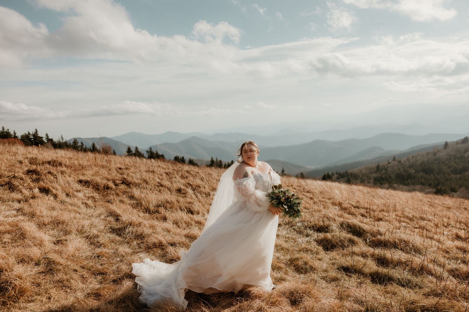 Bride in flowing white gown holding bouquet on a mountain meadow with scenic views.