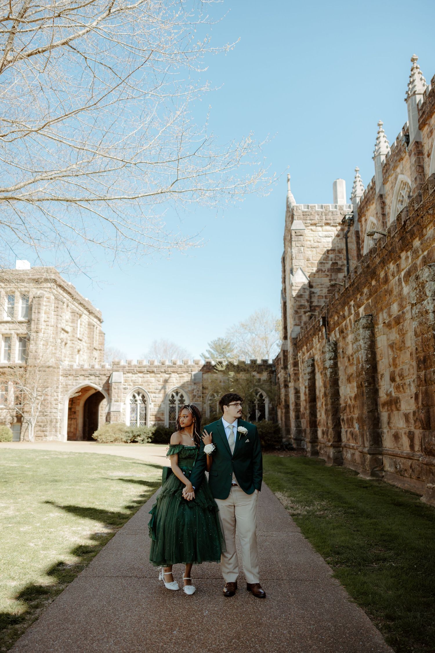 Couple walking hand in hand along pathway beside historic stone castle on sunny day.