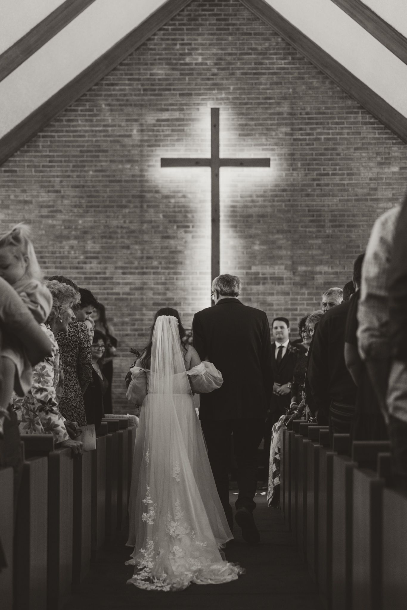A black and white photo of a wedding ceremony inside a church with a prominent cross on the brick wall.