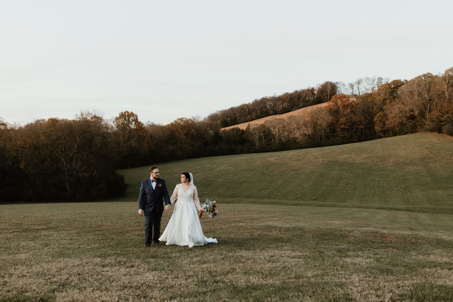 A couple and their dog walk together across a scenic grassy hillside on an autumn afternoon.