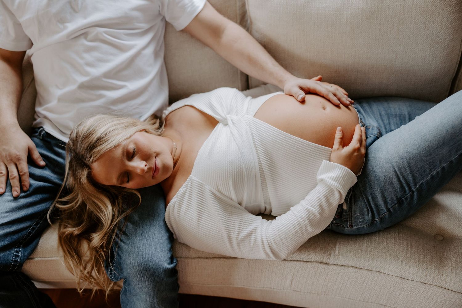 Pregnant woman in white top and jeans resting on a couch, cradling her baby bump while partner sits beside her.