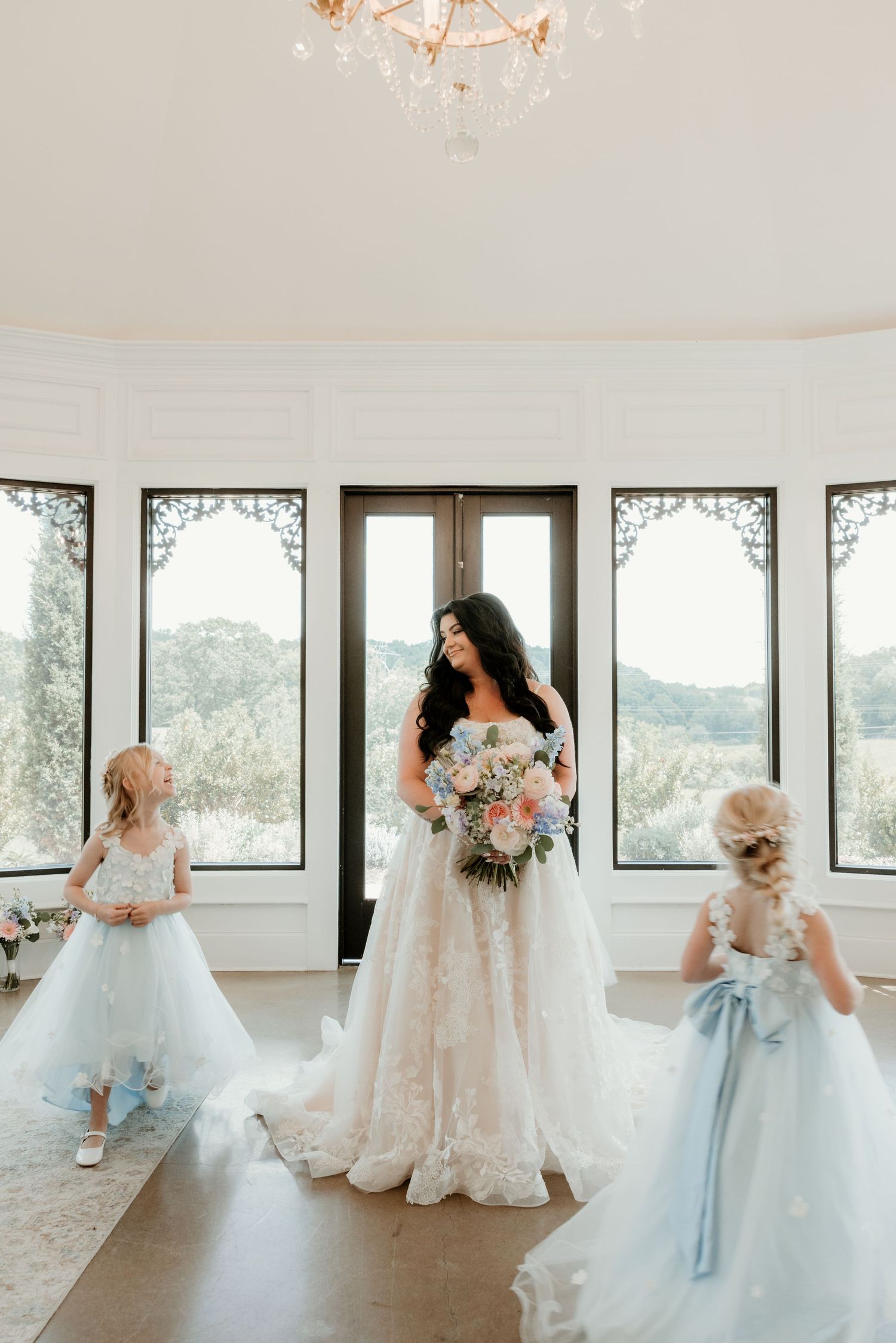 Bride in lace ballgown holds floral bouquet while two flower girls in blue dresses stand beside her.