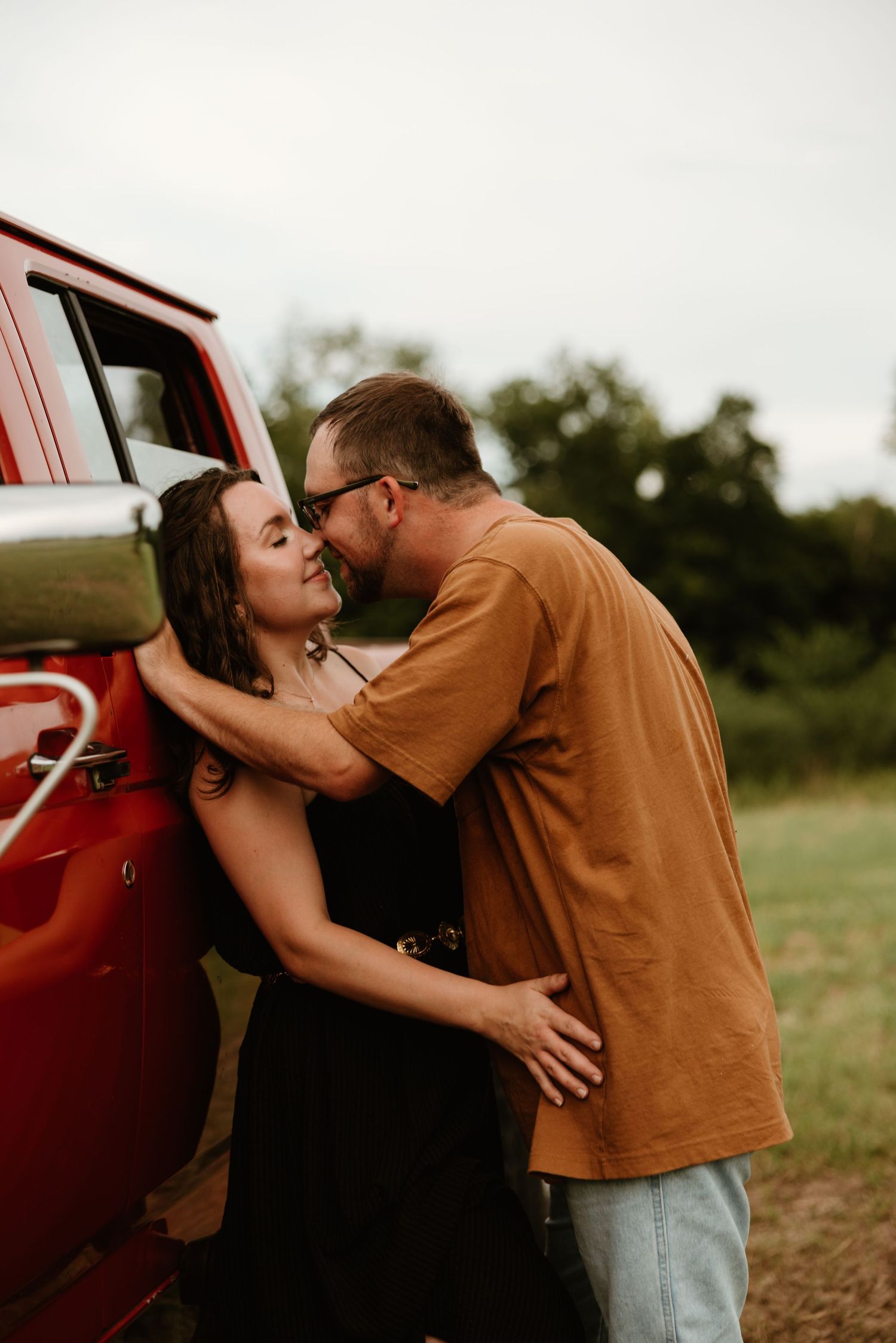 Intimate couple embraces beside a vintage red truck in a rustic outdoor setting.