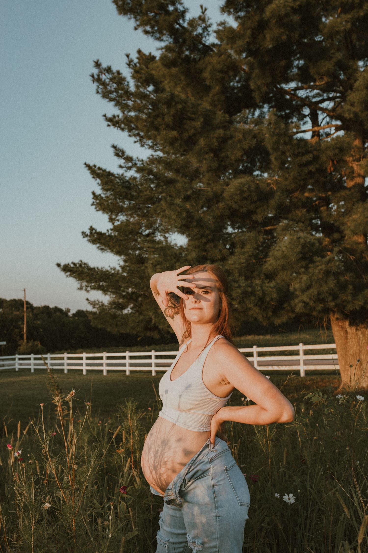 A pregnant person posing outdoors at golden hour in a field with white fencing and pine trees in the background.