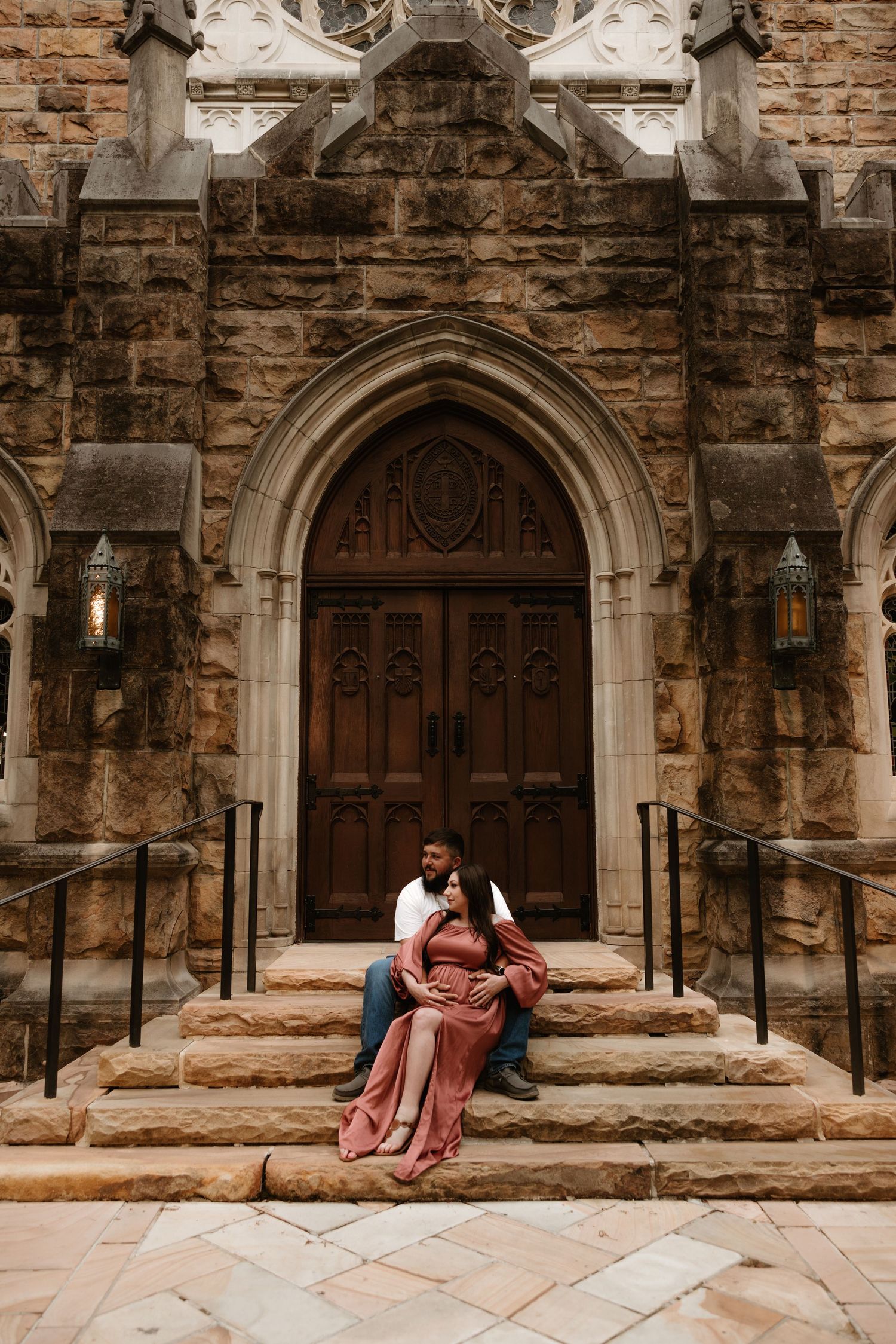 A couple sits on stone steps in front of a gothic church entrance with ornate wooden doors.