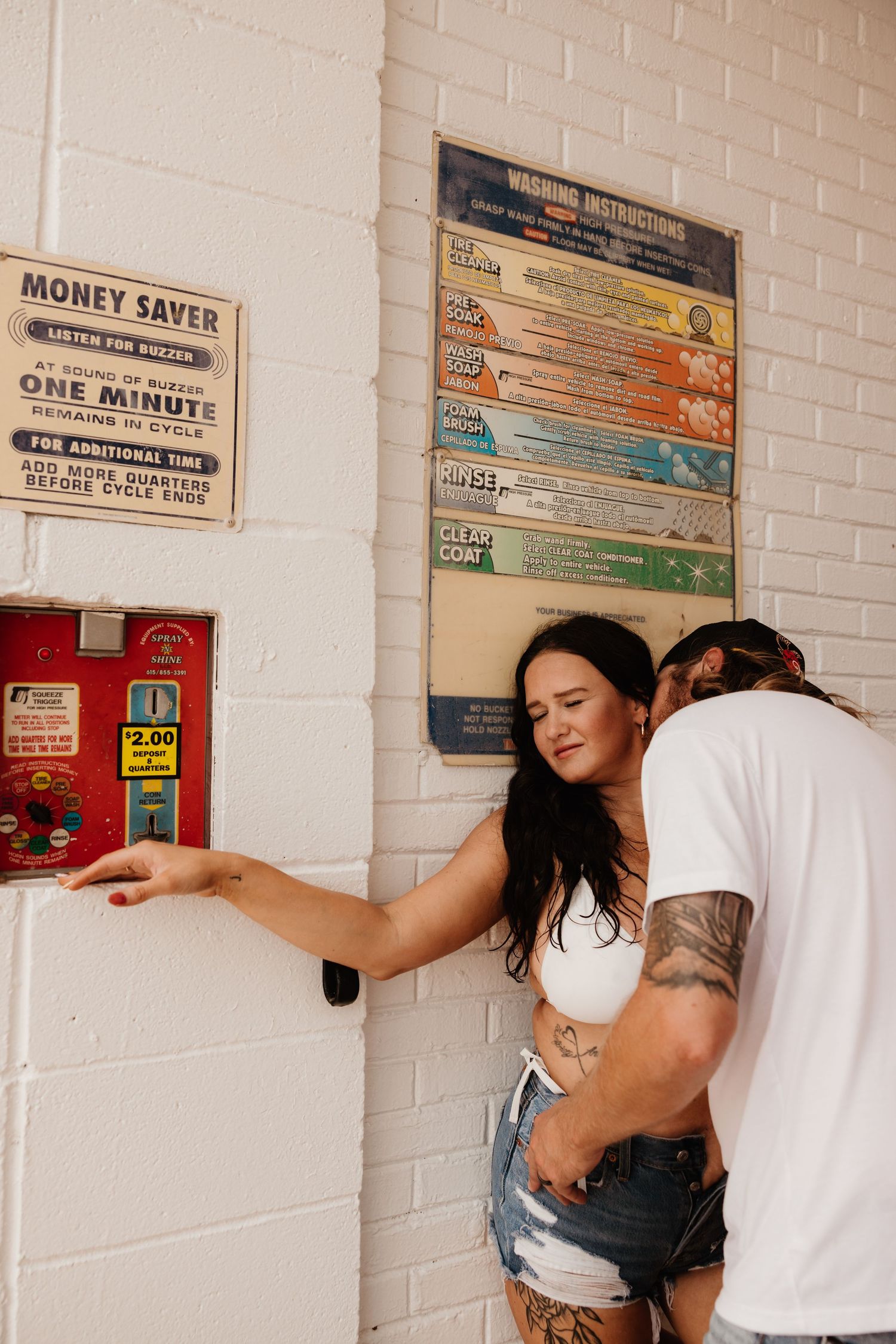 A couple at a car wash pay station with vintage pricing signs on white brick walls.