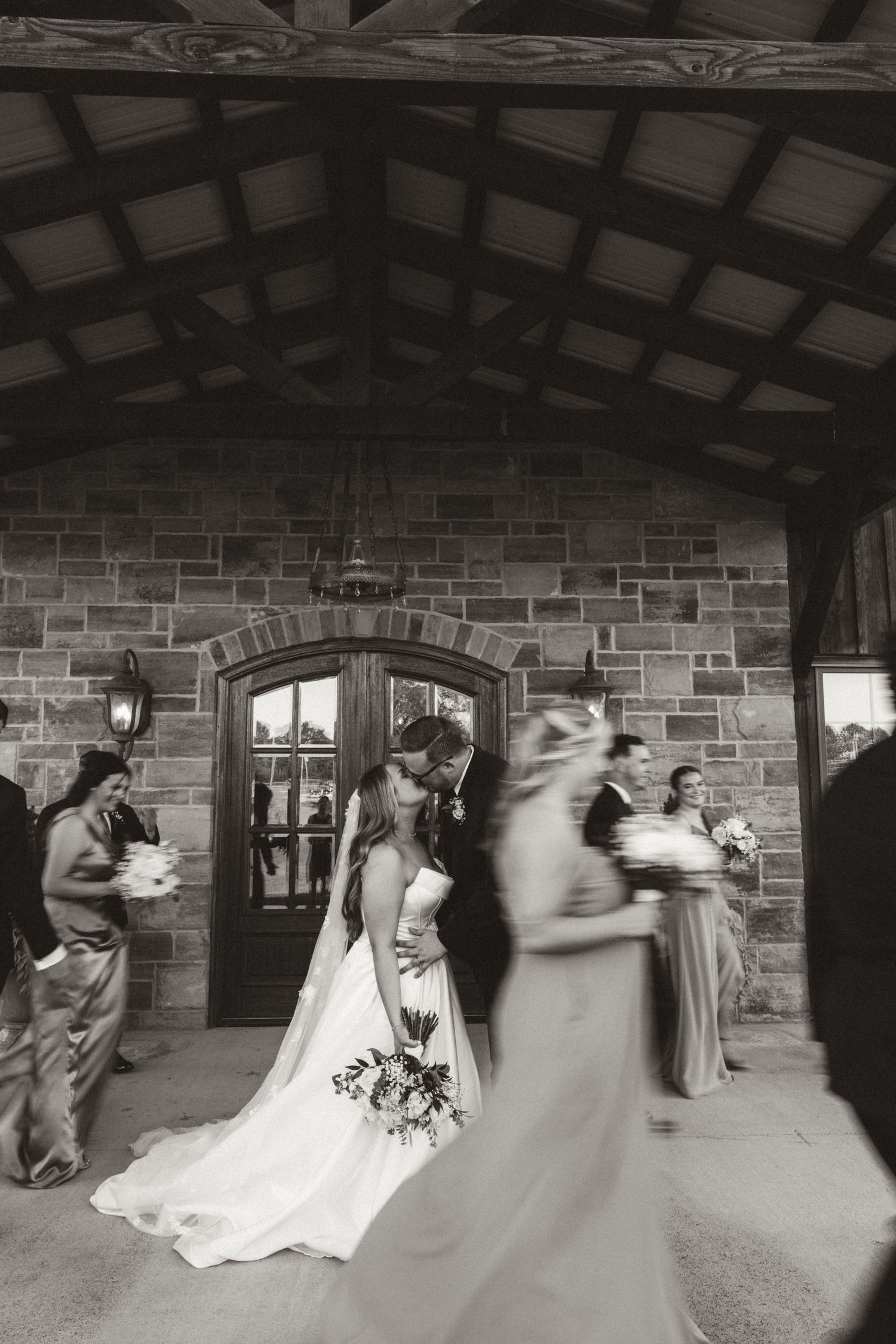 Motion-blurred black and white wedding scene outside stone building with arched doorways and wooden beams.