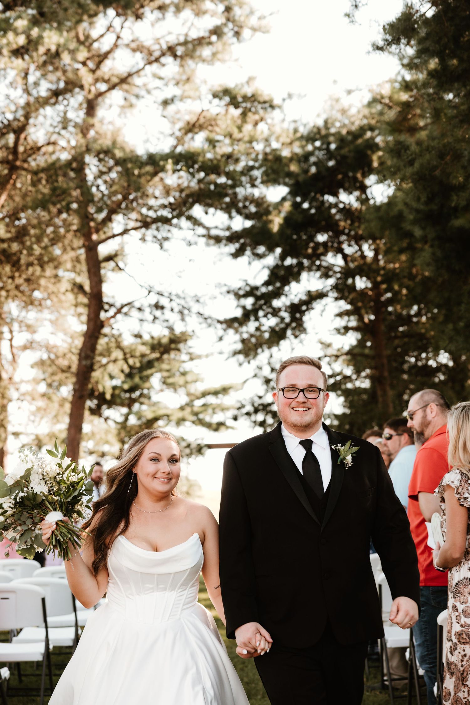 A joyful wedding couple walks together outdoors at sunset with trees in the background.