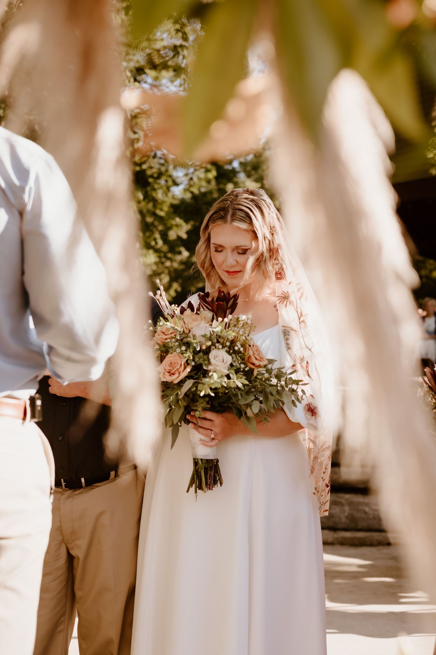 A bride in a white wedding dress holds a romantic bouquet of blush and cream flowers in sunlit outdoor ceremony.