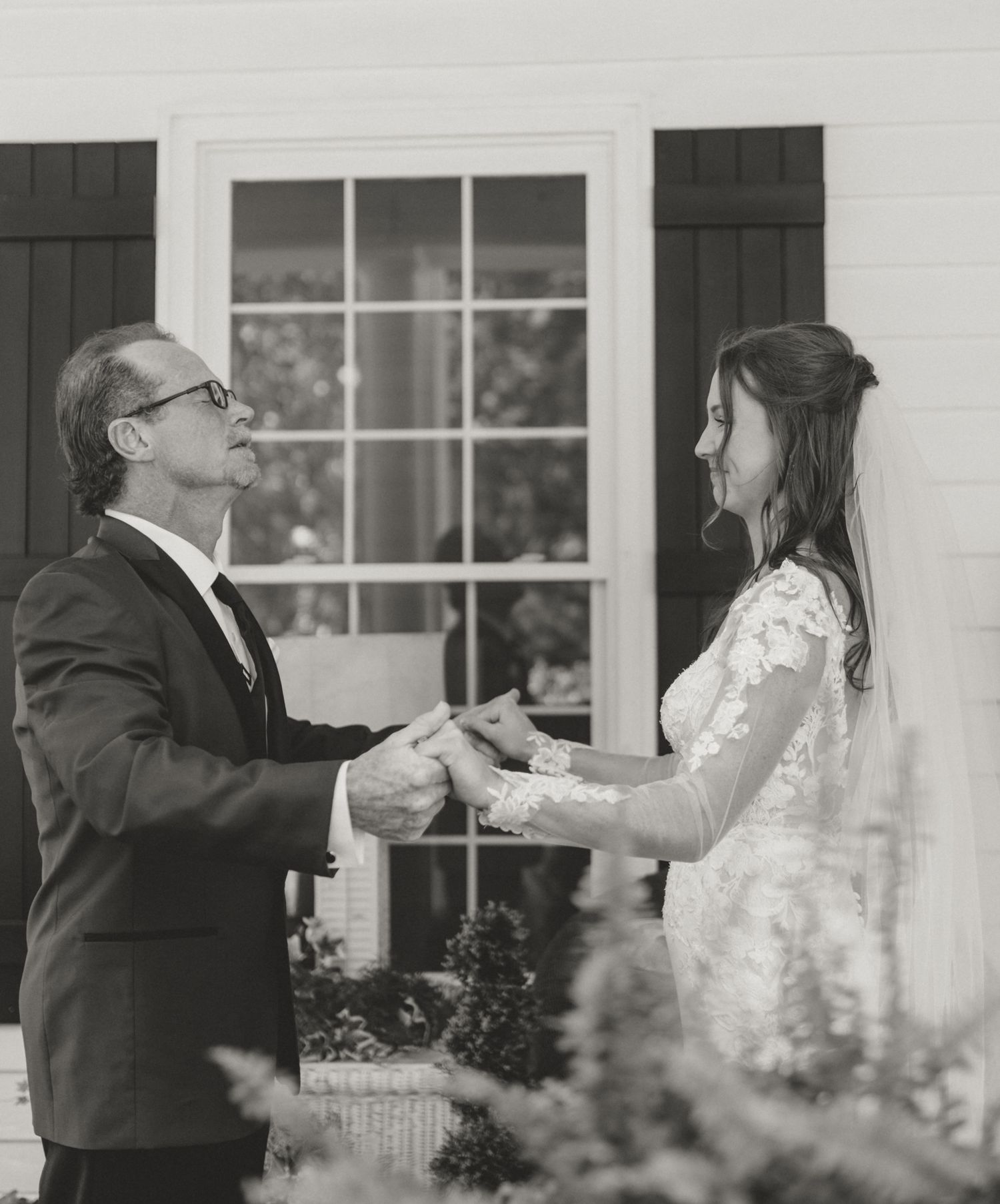 A tender black and white moment shared between two people in formal attire outside a window-lined building.