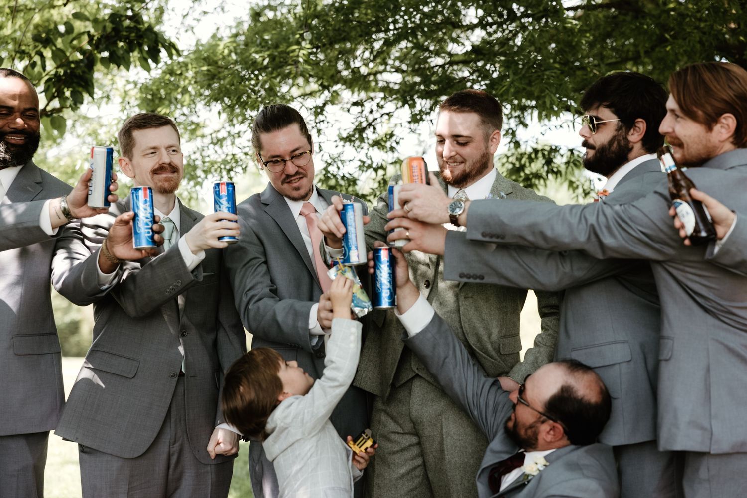 Wedding party in grey suits toasting with beer bottles and cans during outdoor celebration.