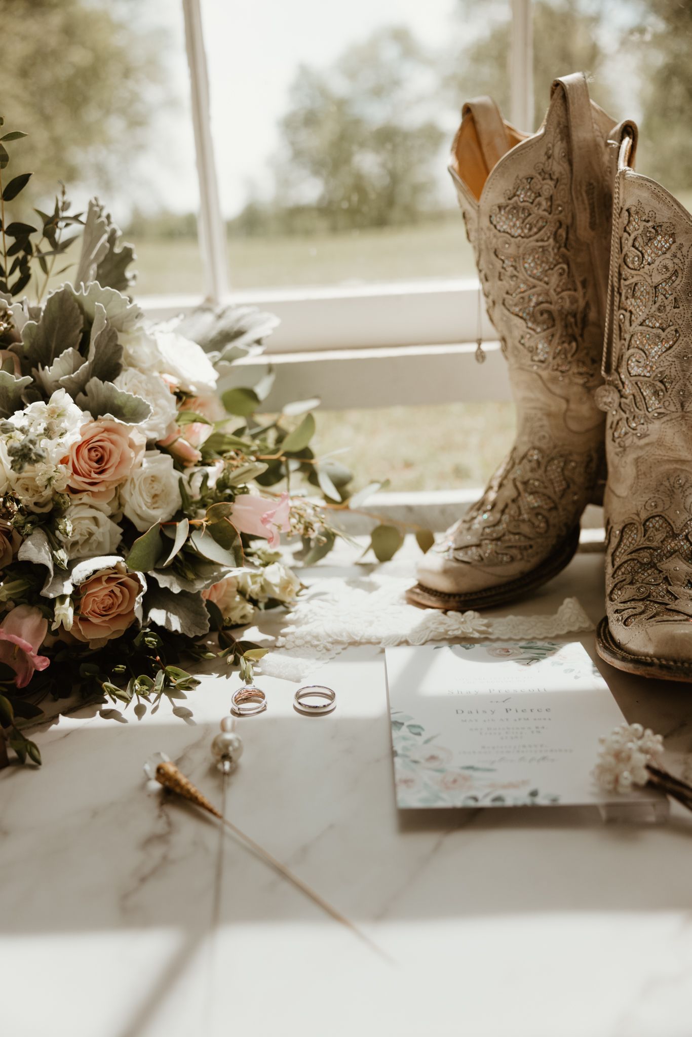 Decorated cowboy boots sit beside delicate floral arrangements and wedding stationery in a rustic wedding detail shot.