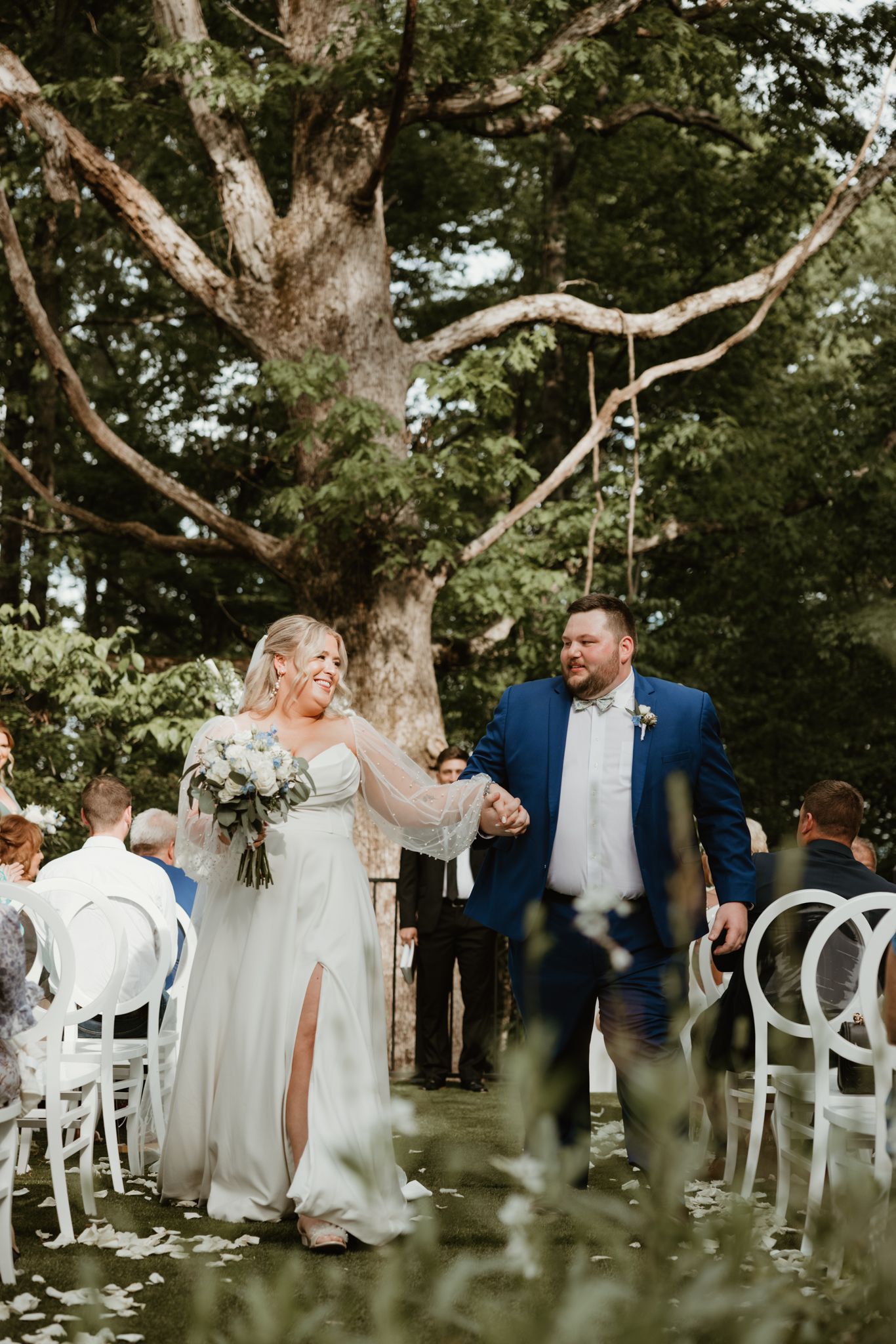 A romantic outdoor wedding ceremony under large trees with white chairs arranged in rows for guests.