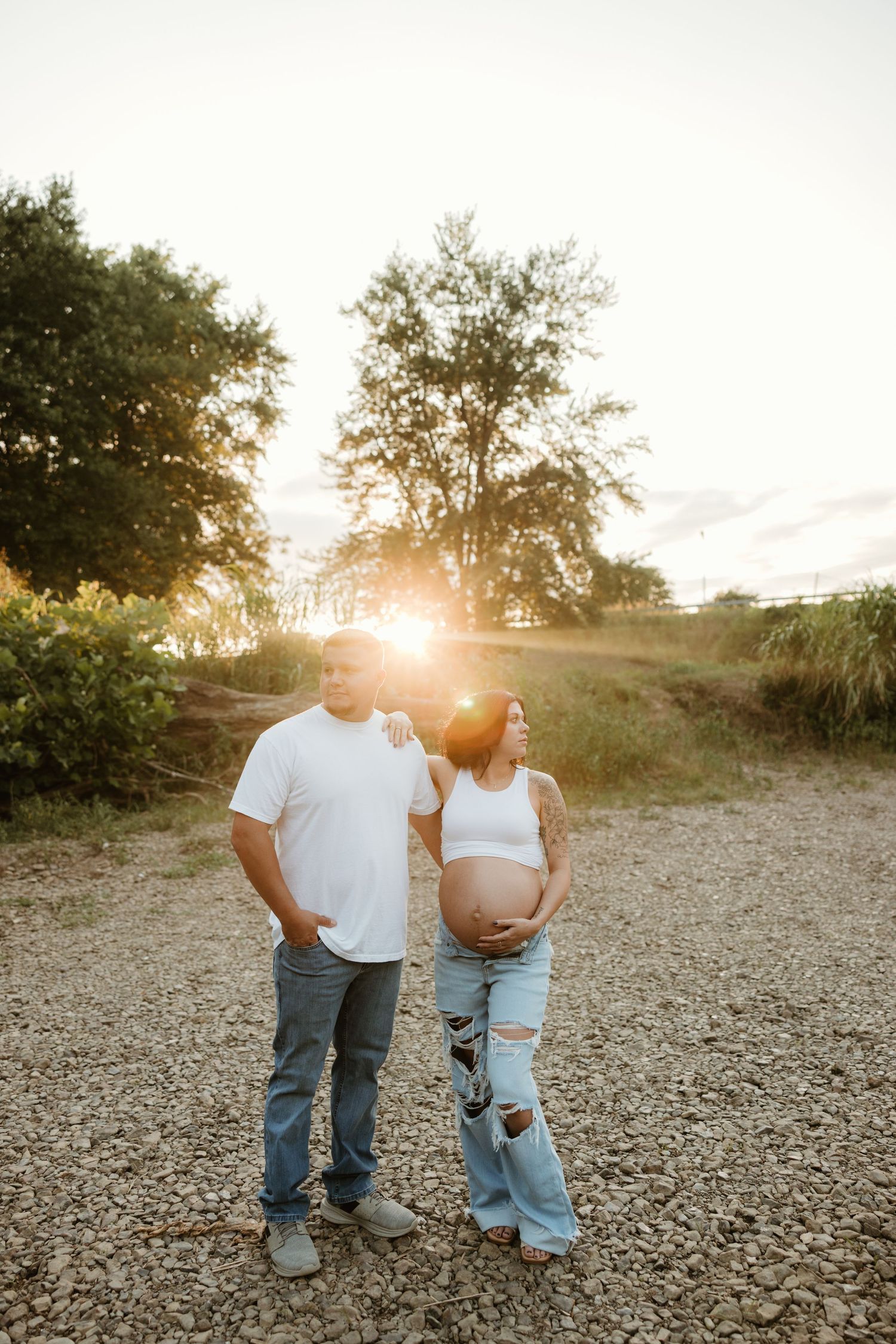 Couple standing together on gravel path during golden sunset with trees silhouetted in background.