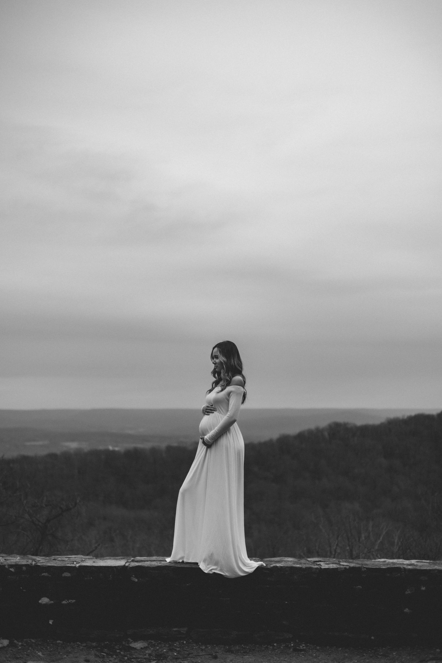 A figure in a flowing white dress stands dramatically on a cliff edge overlooking a misty mountainous landscape.