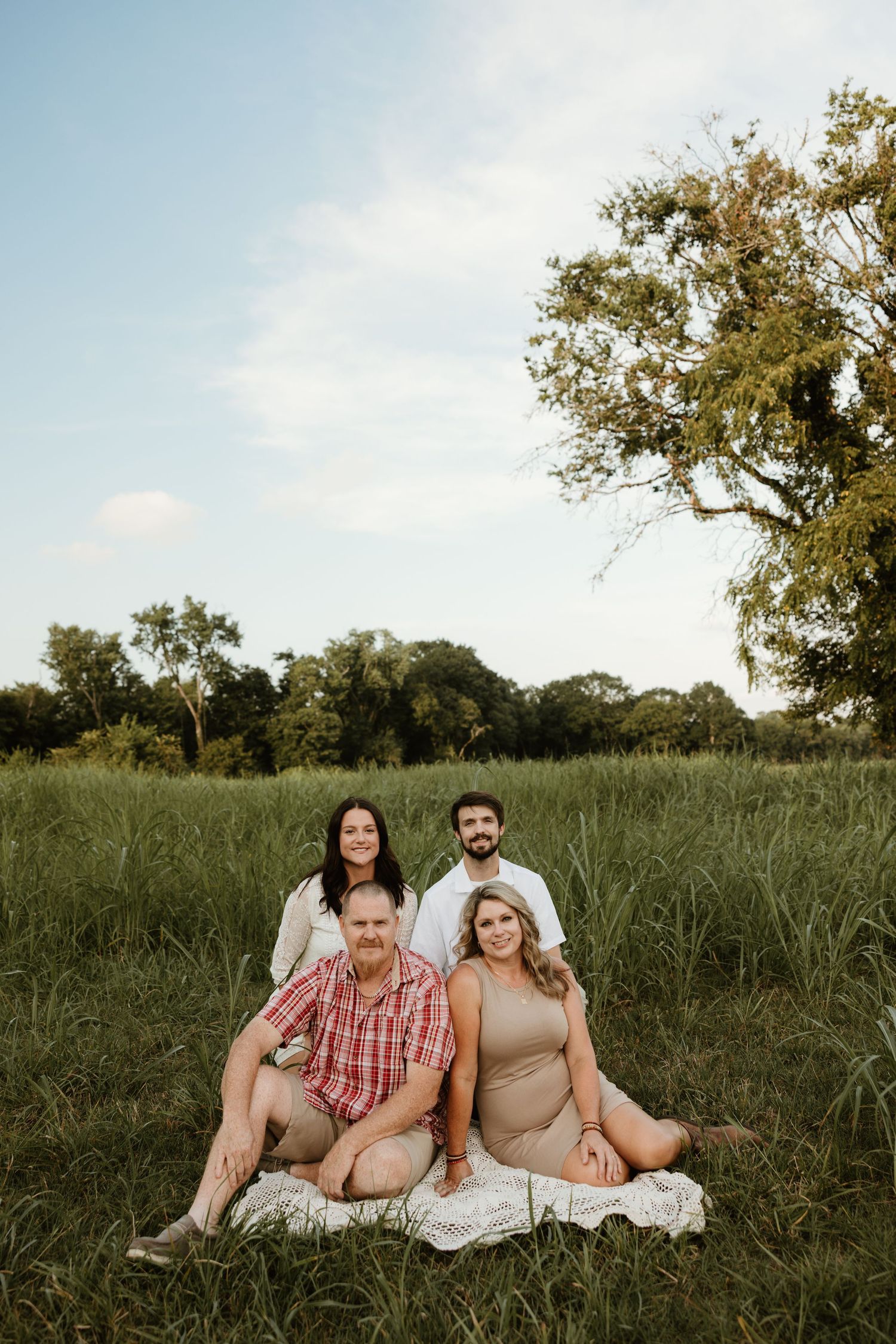 A group lounges together on a white blanket in a grassy field at sunset with trees in the background.
