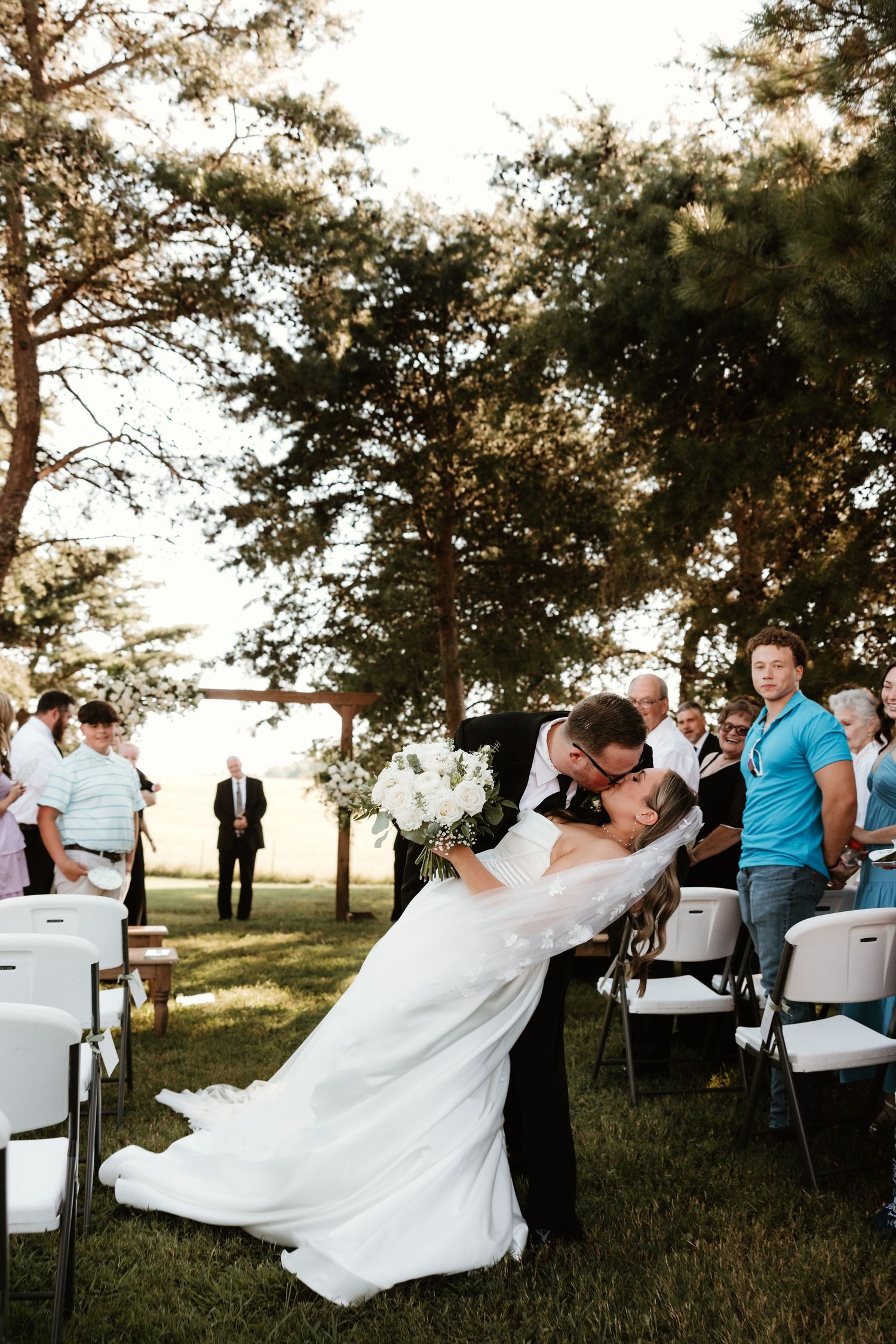 Romantic outdoor wedding ceremony with a dip kiss moment under tall trees at sunset while guests look on from white chairs.