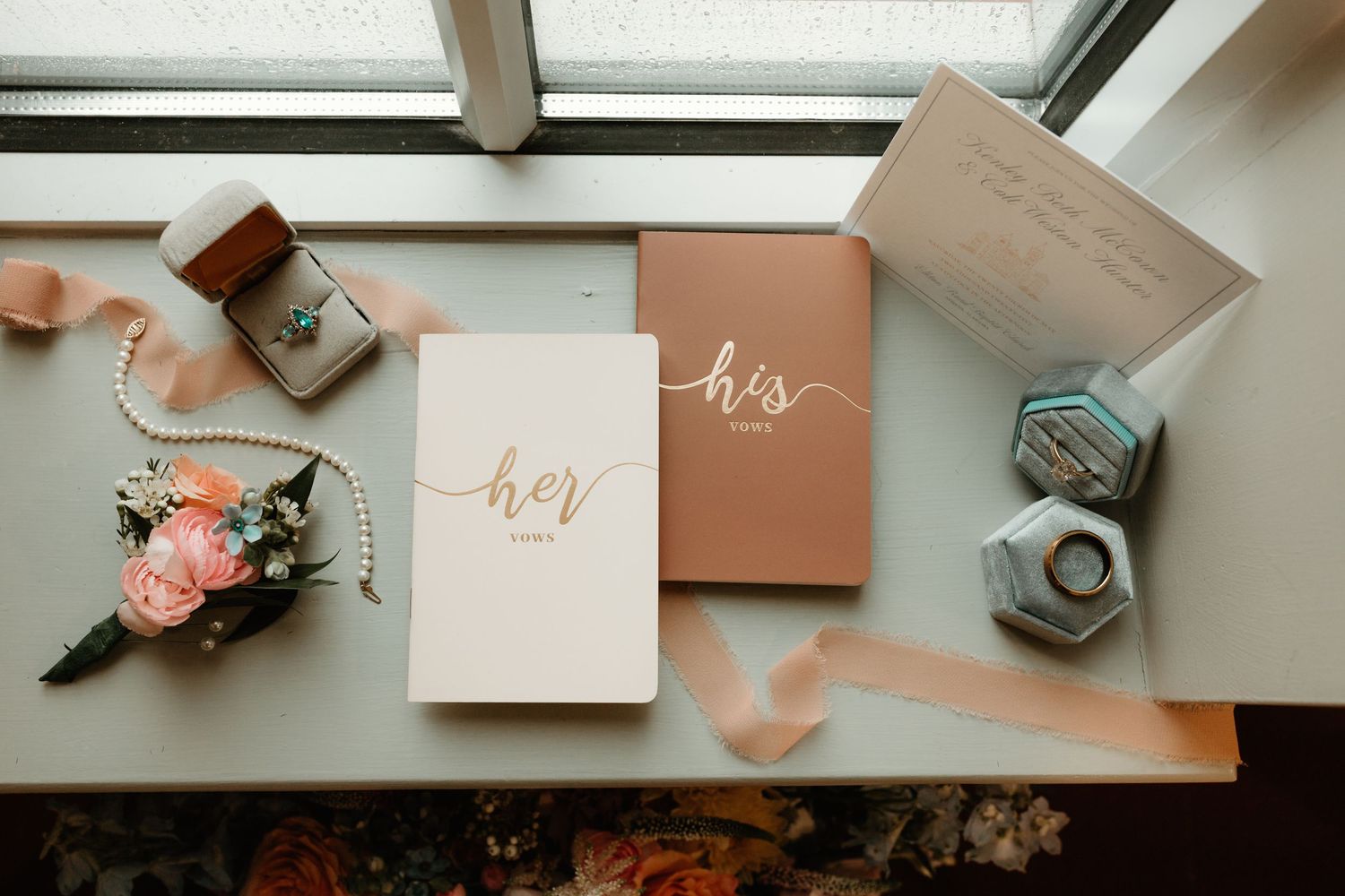 Wedding vows books in rose gold and blush colors with floral arrangements and ring boxes on a white ledge.