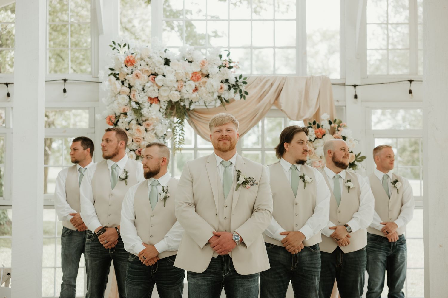 A wedding party in cream-colored suits and jeans poses together in front of a bright glass-walled backdrop.
